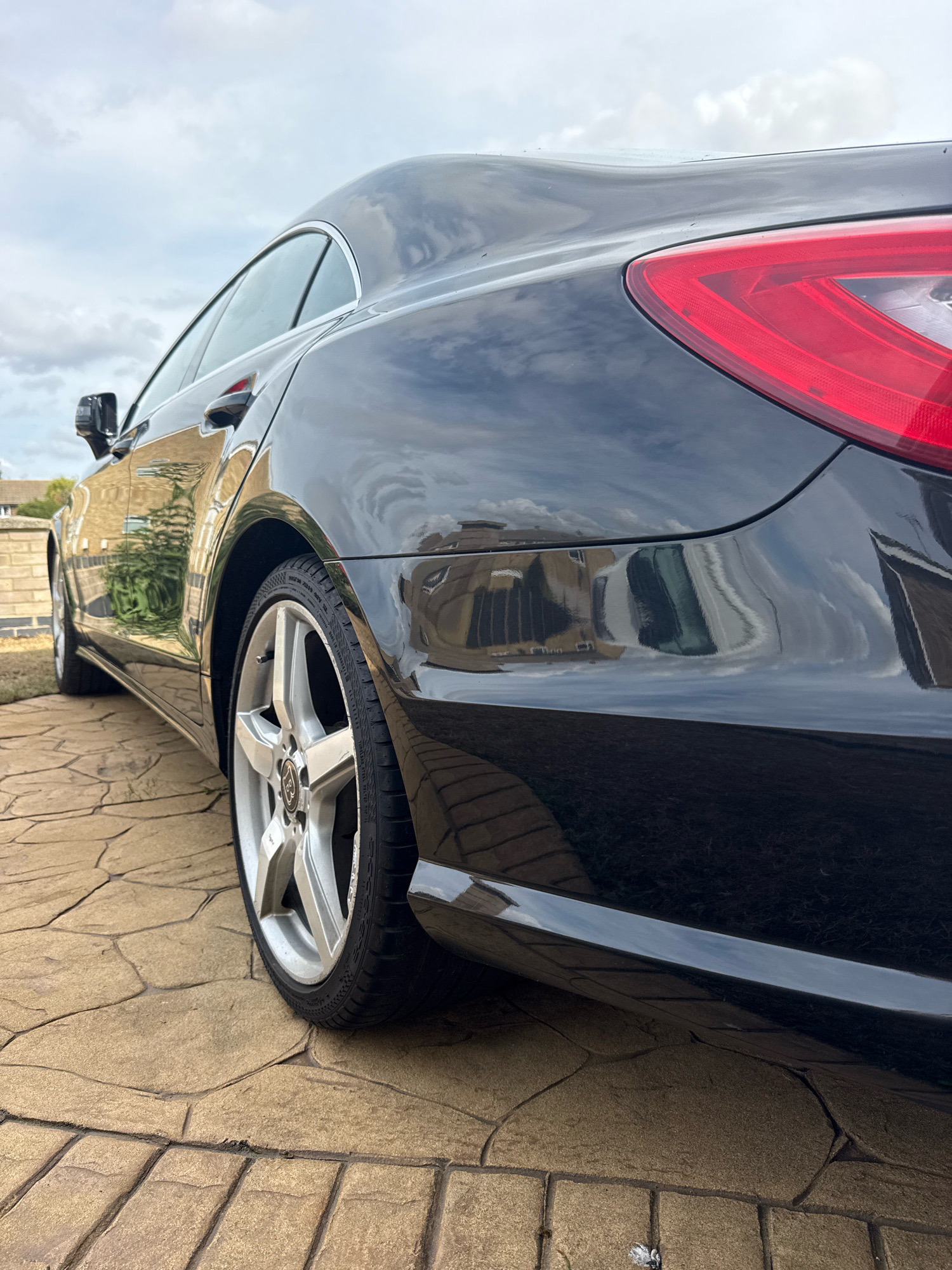 The image shows the rear quarter panel and wheel of a black car parked on a patio of large, irregular-shaped paving stones. The car's paintwork is highly reflective, showing distorted reflections of the sky, clouds, and buildings, including what appears to be a residential property with a brick facade and windows. The car has a distinctive red taillight visible. The setting suggests a residential driveway or garden area in Colchester, United Kingdom. The sky is overcast with visible clouds. No people are present in the image.