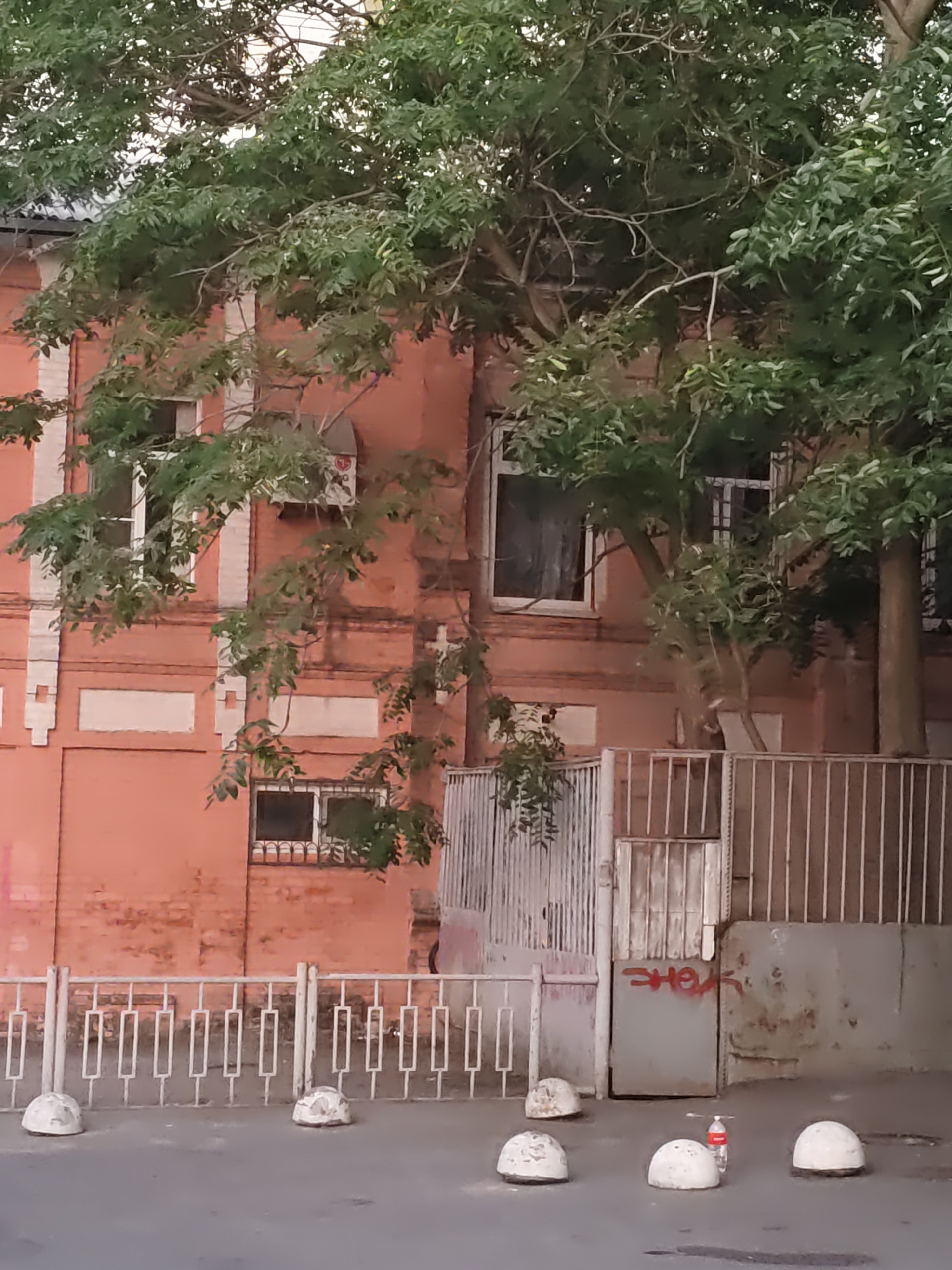 The image depicts a residential building in Rostov-on-Don, Russia. The building has a textured orange facade with white architectural details around windows. Several trees with dense green foliage are growing in front of and around the building, obscuring parts of it. The lower level of the building features a metal gate and a segmented metal fence. In front of the fence, along the street, are several white, dome-shaped concrete bollards. A red-capped plastic bottle is visible next to one of the bollards. Graffiti in red spray paint is present on the metal gate and an adjacent wall. The lighting suggests it is daytime, possibly late afternoon or early evening.