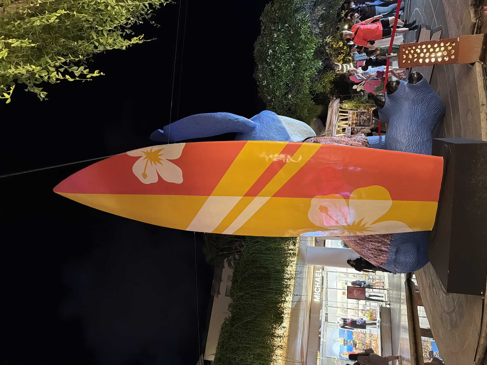 The image captures a nighttime scene in Kuta, Indonesia, likely at Beachwalk Shopping Center. Dominating the foreground is a large, vibrantly colored surfboard sculpture, painted with alternating red and yellow panels and adorned with white hibiscus flowers. The surfboard stands upright, positioned in front of a blue, stylized sculpture that appears to be an abstract representation of a creature, possibly Stitch from Disney's Lilo & Stitch, given its blue color and large ears. A person, partially obscured by the surfboard, is visible behind it. In the background, the facade of a shopping center is illuminated, with storefronts including "MICHAEL KORS." Several people are browsing or walking in the shopping area and surrounding plaza. The environment is an outdoor plaza with water features and lush greenery, including hanging vines and trees, illuminated by decorative lights. The overall atmosphere is that of a lively, commercial area at night.