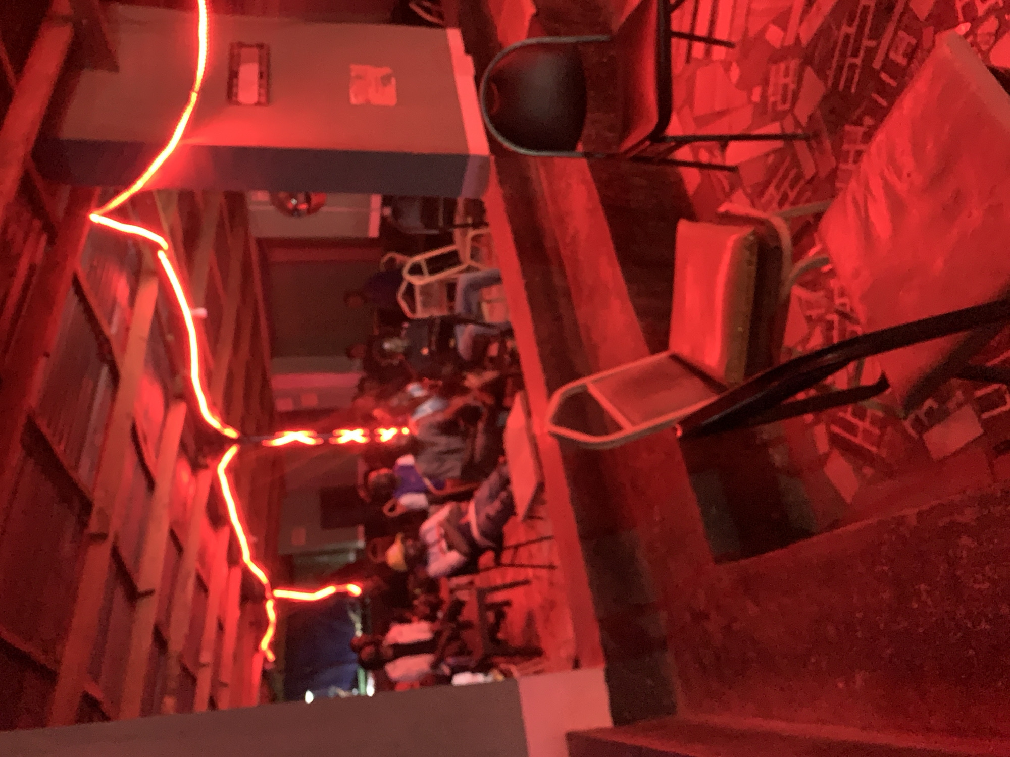 The image depicts a room interior with a group of people seated at tables and chairs. The room is illuminated by red lighting, possibly neon or LED strips, which gives a strong red cast to the scene. The walls on the left side appear to be lined with panels or stacked shelves. Tables and chairs are scattered throughout the space. Individuals are seated at some of the tables. Based on the prompt, the location is New Stadium, Nigeria.