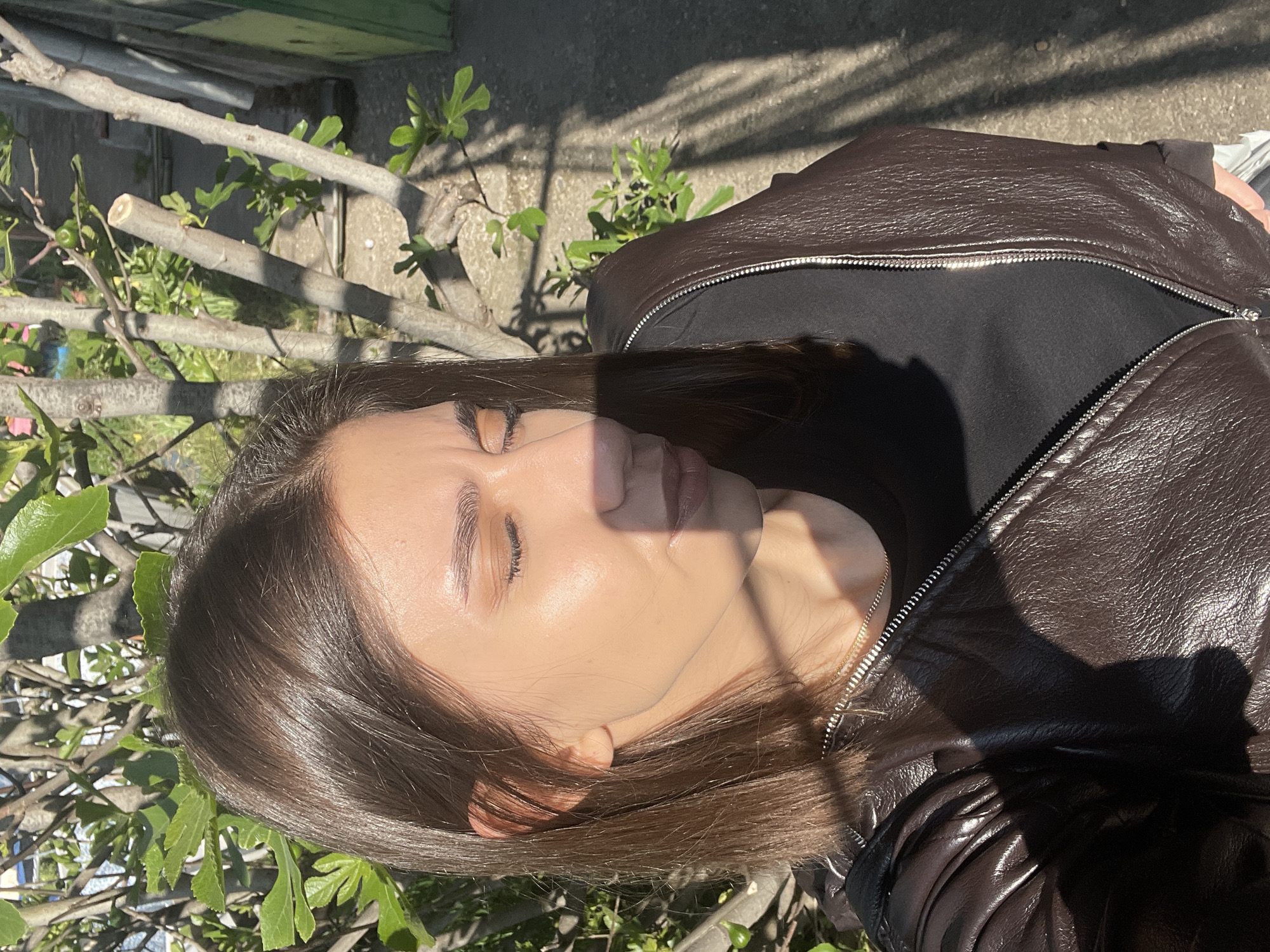 A medium-shot photograph shows a woman with long brown hair, fair skin, and closed eyes. She is wearing a black top and a brown leather jacket with a partially open silver zipper. Her face is partially covered in shadow. The background includes green foliage and branches, with a glimpse of a concrete surface. It is impossible to know the exact location in Sofia, Bulgaria from the given image.