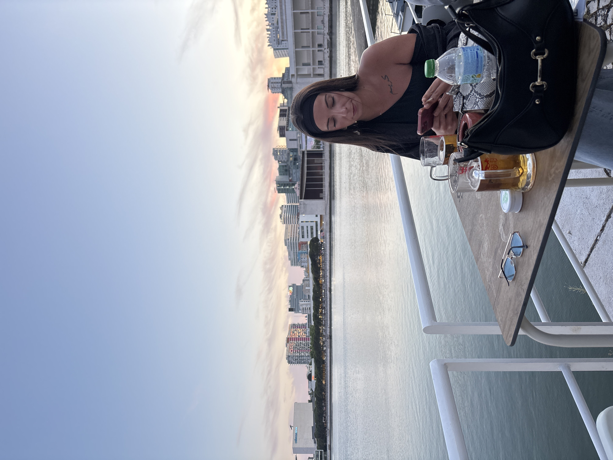 A woman is seated at a table on what appears to be a boat or ferry, with a cityscape visible in the background, likely Lisbon, Portugal, based on architectural styles. The cityscape is lit with a sunset with pink and gold hues. The woman has dark hair and is wearing a black top and a black headband. She appears to be looking at her phone. On the table, there are glasses of beer, a bottle of water, sunglasses, a container of mints and a black purse. The background shows a body of water adjacent to the city's buildings.