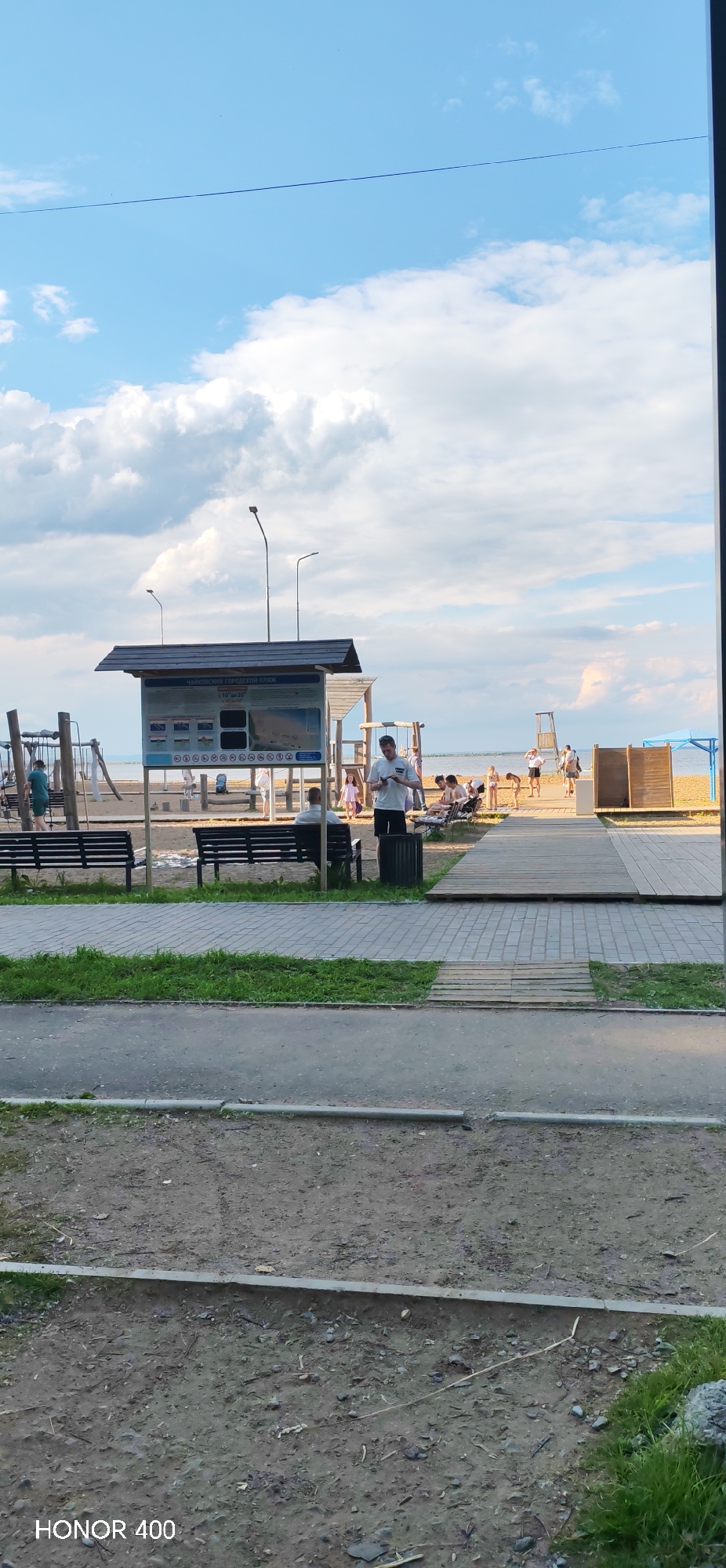 The image depicts a view of a city beach in Chaikovsky, Russia. The beach is sandy with scattered people enjoying the area. In the foreground, there are paved pathways and grassy areas, with benches placed for seating. Behind the benches, a wooden structure with a roof serves as an information board, displaying maps and regulations for the beach. Various individuals are visible on the beach, some sitting on benches, others walking or standing. Children's play equipment, such as swings and climbing frames, is also present on the sandy area. Further in the background, the calm water of a body of water can be seen, with a blue sky dotted with white clouds above. The overall scene suggests a recreational public space on a pleasant day.