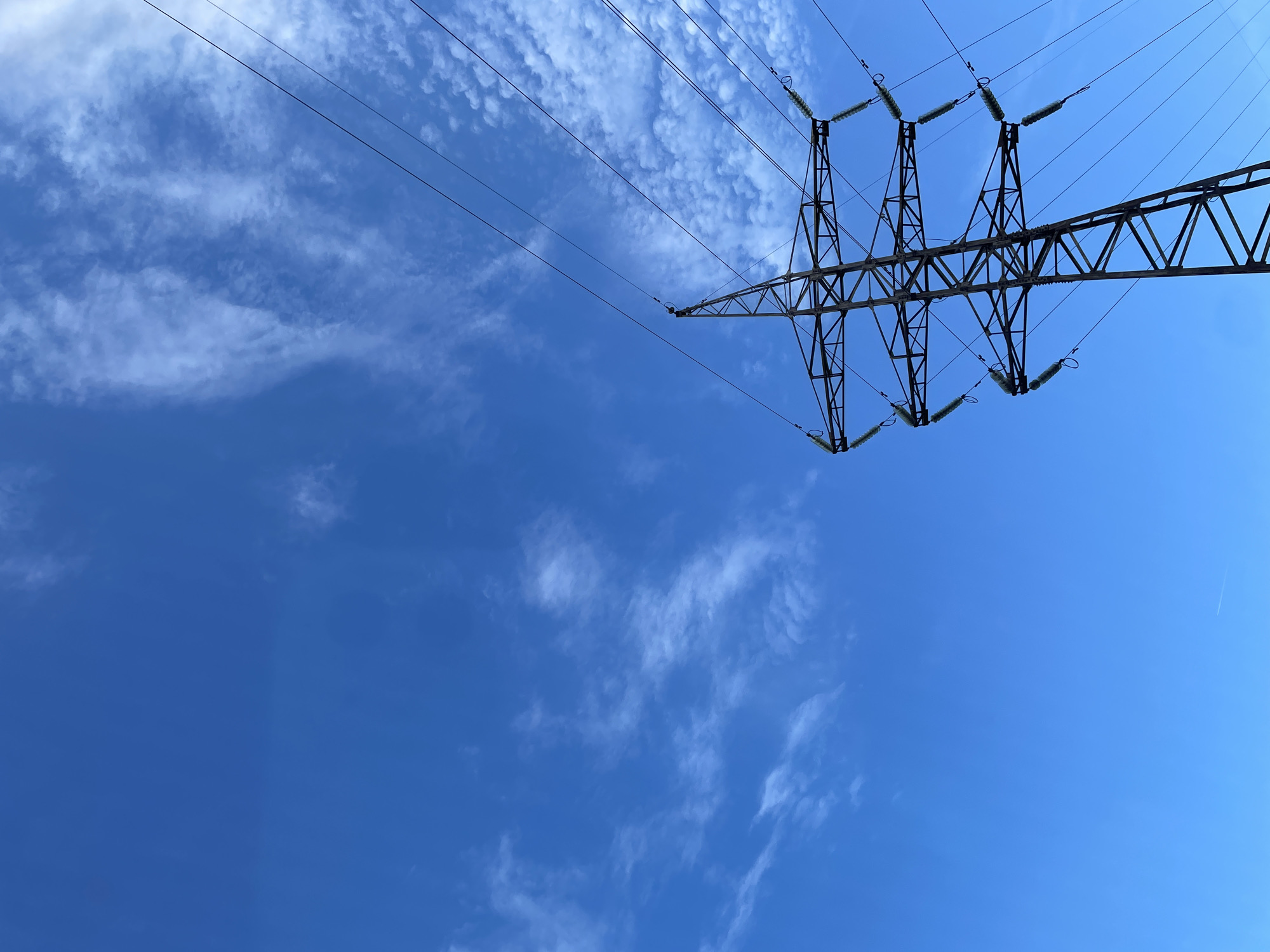The image depicts the upper and mid-section of a metal lattice electrical transmission tower, commonly known as a pylon, set against a bright blue sky with scattered white, wispy clouds. The tower's structure is visible, comprising multiple interconnected steel beams forming a robust framework. Several sets of green or teal-colored glass/ceramic insulators are suspended from the tower's cross arms, supporting at least six high-voltage power lines that extend out of the frame towards the upper left and right. The power lines appear taut and dark against the sky. No individuals, vehicles, or specific ground features are visible, as the perspective is upward-angled. The lighting indicates daylight conditions. The specific location within the city of Izhevsk, Russia, is not discernible from the visual content of the image.