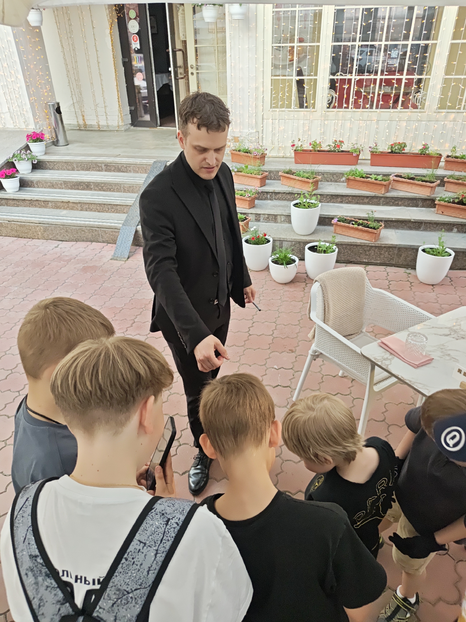 The image shows a magician performing for a group of young boys in an outdoor setting. The magician, dressed in a black suit, is standing and gesturing with a small object in his right hand. The boys are gathered around him, looking up at the magician. One boy is holding a cell phone, possibly recording the performance. The setting appears to be a patio or outdoor dining area of a restaurant. There are flower pots and plants on the steps behind the magician. The patio is paved with red patterned tiles. A white table and chairs are to the right of the group. The location is Yekaterinburg, Russia, as indicated by the "Я" logo on a sign visible through the window in the background.