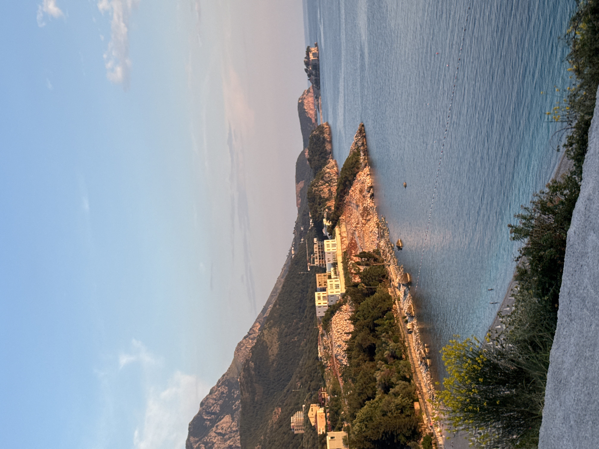 The image shows a coastal scene in Kamenovo, Montenegro. A rocky peninsula juts into the blue sea. Several modern white buildings are nestled along the hillside of the peninsula. On the left, a larger mountain rises, with some residential buildings visible on its lower slopes. A thin beach runs along the base of the peninsula. In the sea, a line of buoys stretches across the water. The sky is light blue with some light clouds. The foreground shows a rock face with some greenery.