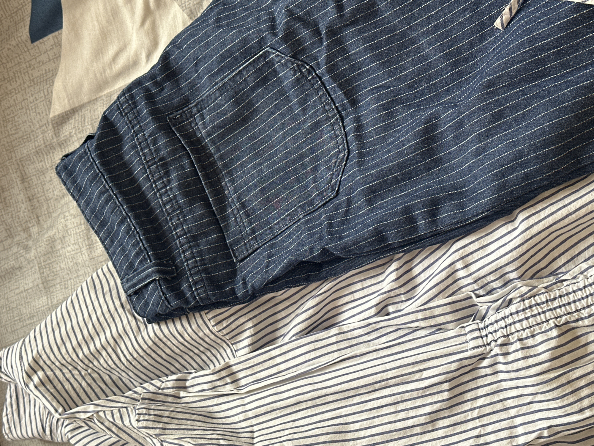 The image shows a close-up of clothing items on a bed. A pair of dark blue, striped shorts with a visible pocket are on top of a white and blue striped shirt. The background consists of a patterned bedspread. There is no indication of the city of Agarak, Armenia, or any other location information in the image.