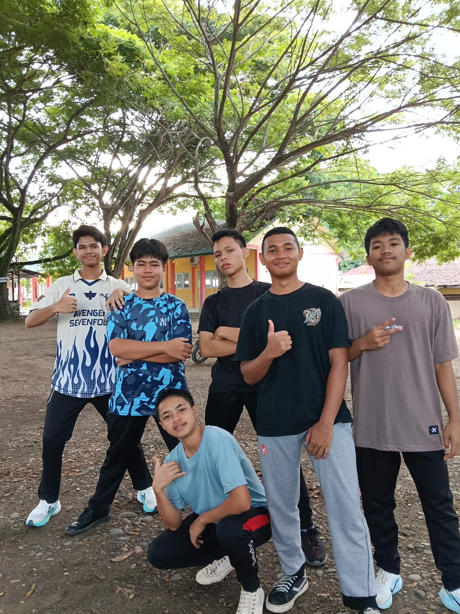 A group of six young males poses outdoors, under the shade of large trees. The setting appears to be a park or open space near a building with a yellowish facade and red accents. The ground is dirt and gravel. The males are dressed in casual clothing, including t-shirts, track pants, and sneakers. Several of them are giving a thumbs-up gesture. One male is squatting. The location is indicated as Lhokseumawe, Indonesia.