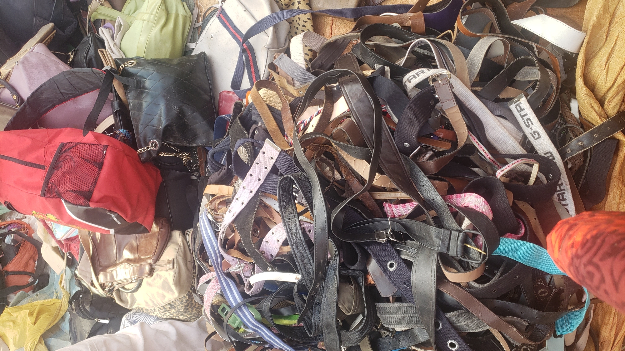 A pile of assorted belts and bags is displayed outdoors in what appears to be a market setting in Kakuri Gwari, Nigeria. The foreground is dominated by a jumbled mass of belts, made from various materials including leather and fabric, in an array of colors and widths, some with buckles and others with eyelets. Mixed in with the belts are several handbags, including a prominent red backpack with black trim and mesh pockets, a quilted black handbag with a gold chain strap, and other bags in shades of beige, brown, and green. Some of the bags exhibit designer logos or patterns. The ground beneath the pile is covered in a rough, textured fabric, likely a tarp or ground cloth. The lighting suggests it is daytime.