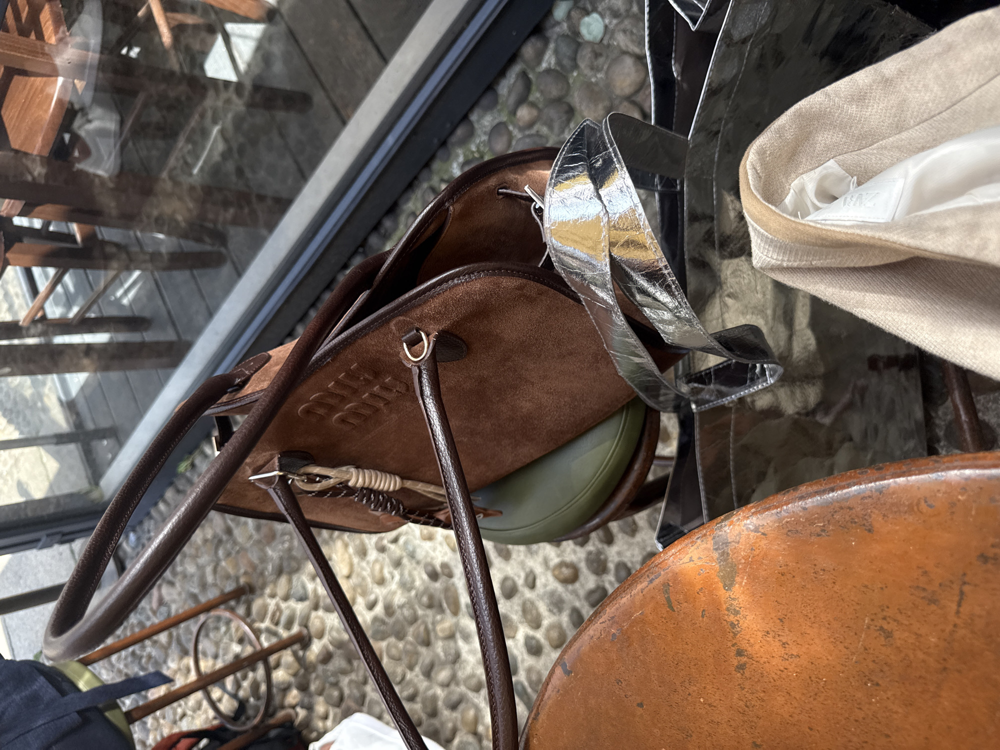 This overhead shot depicts personal belongings in an outdoor or semi-outdoor setting, likely a café terrace in Milan, Italy.
The main subject is a brown suede handbag with dark brown leather handles and trim. The brand name "MIUMIU" is embossed on the front of the bag, though partially obscured by a handle. Inside, a glimpse of an olive green object is visible. Next to the brown bag is a prominent silver accessory, possibly another bag or a wide strap, featuring a distinctive cracked metallic texture.
In the upper right corner, a light beige knitted garment, possibly a sweater or cardigan, is visible. An internal white label clearly displays the brand "ZARA."
The ground beneath the objects consists of small, rounded cobblestones or pebbles, visible directly and through reflections in what appears to be a glass surface. These reflections also show abstract shapes and hints of wooden furniture, suggesting tables or chairs. A weathered, coppery-orange surface, likely a tabletop, occupies the bottom right of the frame. The scene is well-lit by bright natural daylight.