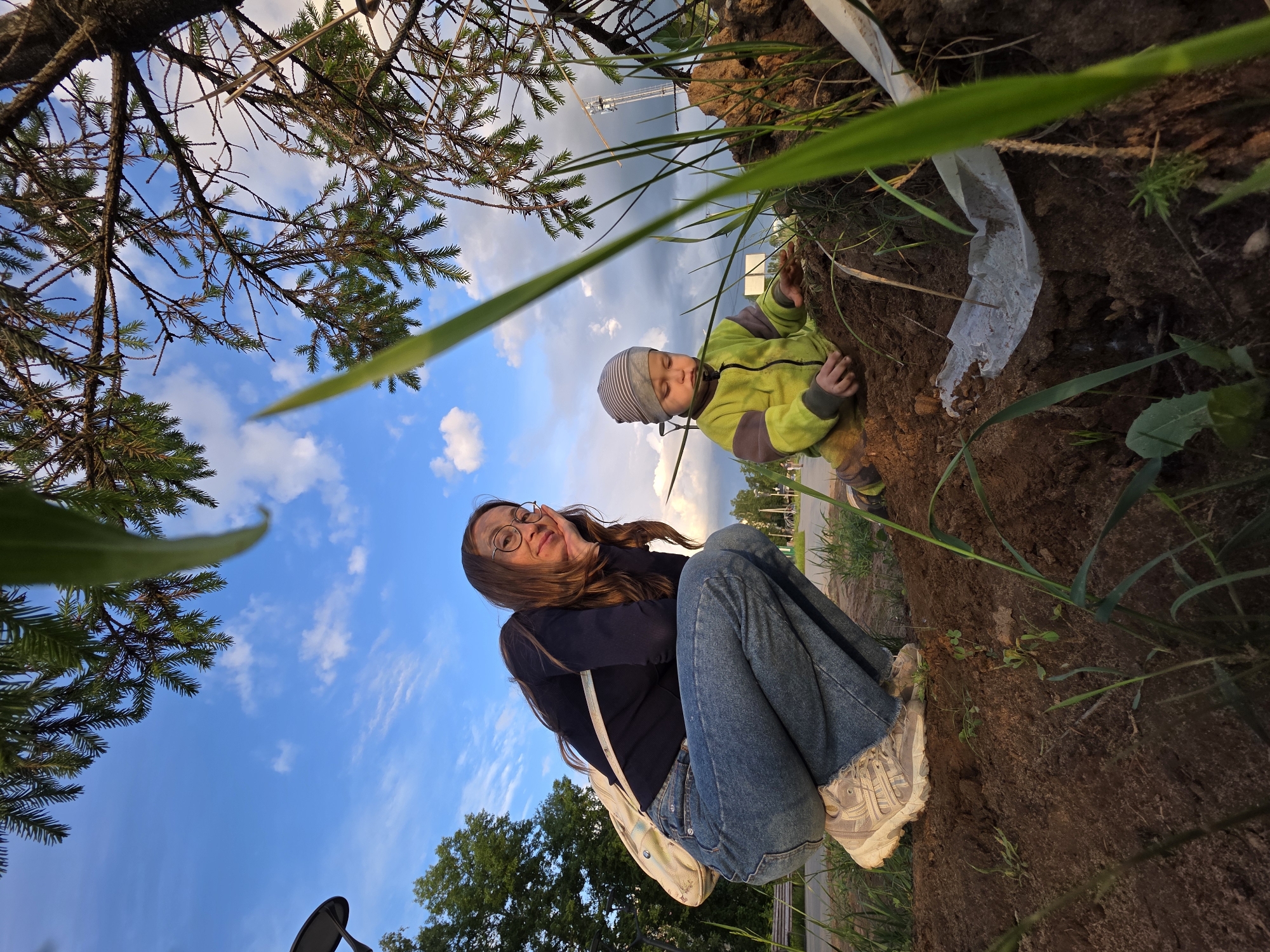 In Chaikovsky, Russia, a woman with long brown hair, wearing glasses, a dark top, jeans, and sneakers, is sitting on an embankment next to a child in a yellow jacket and striped hat who is partially buried in the dirt. The scene is viewed from a low angle, looking up at the subjects against a blue sky with scattered clouds and a foreground of tree branches and grass. A white plastic sheet lies near the child in the dirt. A park with trees and benches is visible in the background.