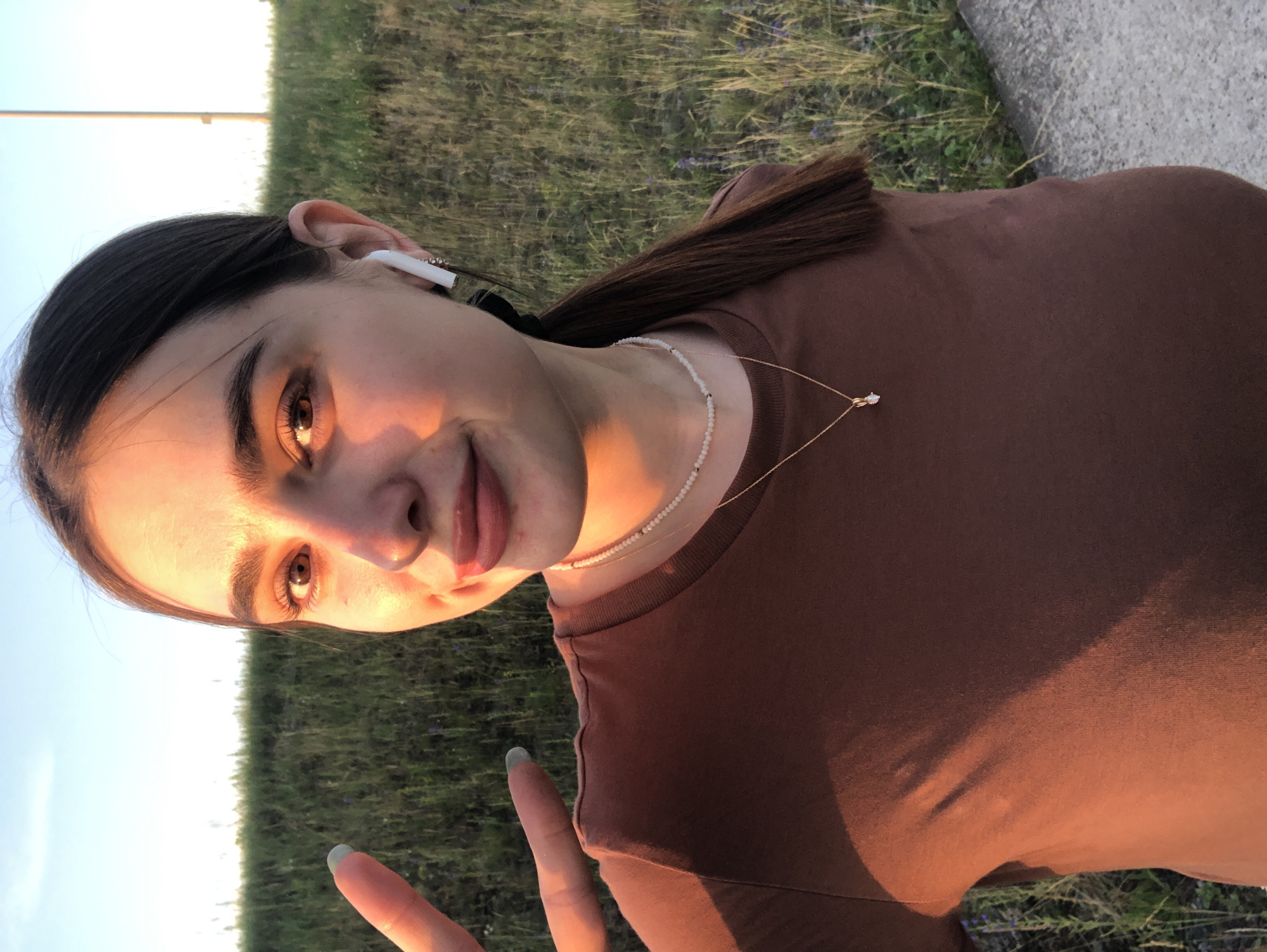 The image captures a young woman outdoors during what appears to be golden hour, indicated by the warm, soft lighting casting long shadows and illuminating her face. She is positioned in a way that suggests she is taking a selfie, with the camera angled upwards towards her. The woman has dark hair pulled back, is wearing white wireless earbuds, and has a delicate pearl necklace and a thin gold chain with a small pendant. She is dressed in a brown t-shirt. Her expression is relaxed and pleasant, with a slight smile, and she is making a peace sign with her right hand, which has a light grey nail polish.
The background consists of a grassy field with hints of greenery and possibly some wildflowers, suggesting a rural or park-like setting in Noviy, Russia. A paved surface, likely a road or walkway, is visible on the right side of the frame. The sky above is a clear, light blue, further confirming the outdoor, daytime setting. The overall mood of the image is serene and casual, capturing a moment of personal enjoyment in nature. There is no visible text in the image.