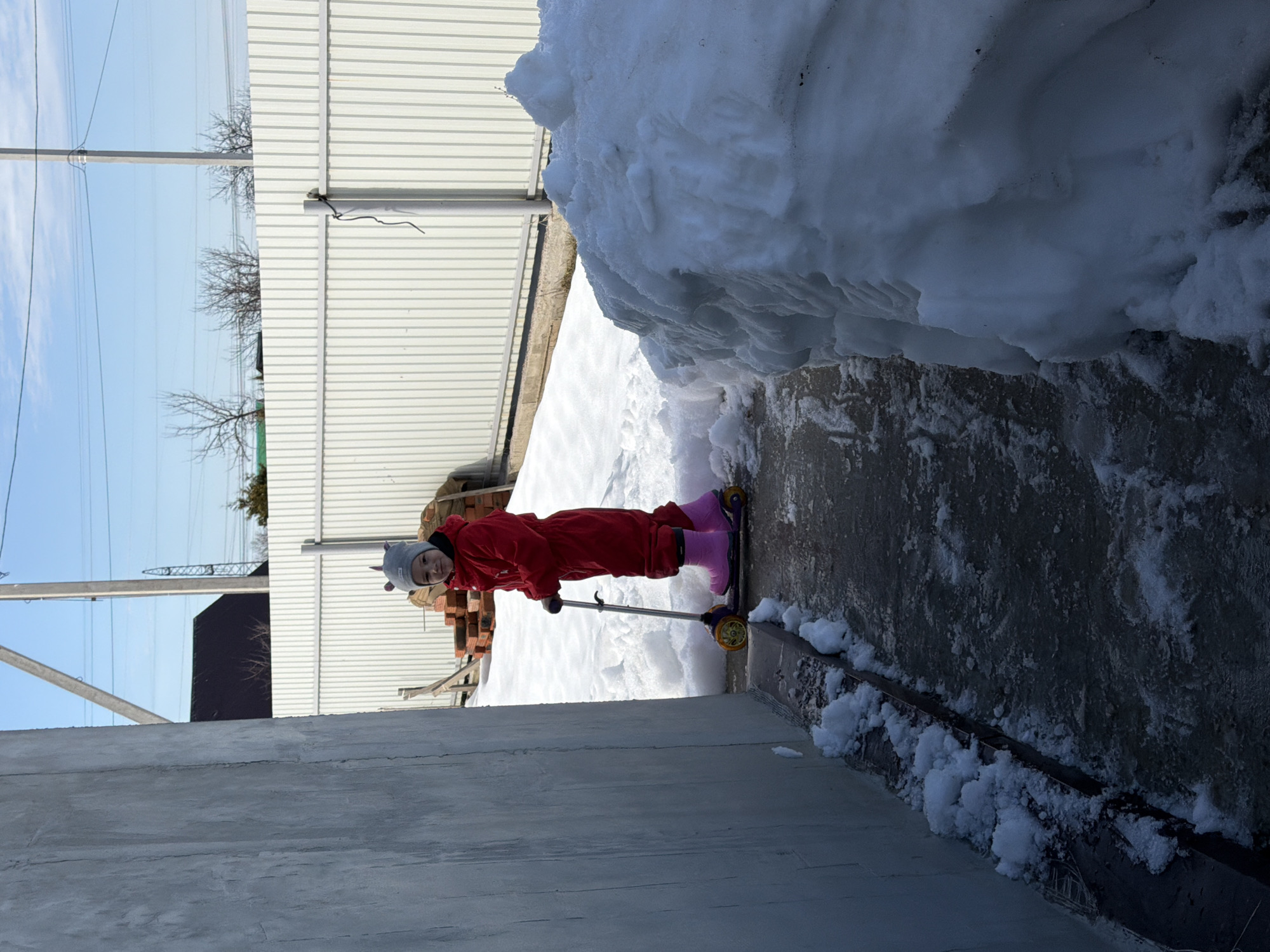 In Chaikovsky, Russia, an outdoor scene depicts a young child on a partially cleared path during winter or early spring conditions. The child, dressed in a red full-body snowsuit, a grey hat with unicorn-like ears, and pink winter boots, stands holding the handlebars of a purple and yellow three-wheeled scooter. The child looks directly forward, towards the camera.
To the immediate left, a tall, textured grey concrete wall borders the path, with small accumulations of snow and ice along its base. The path itself is wet and appears icy in places. To the right, a large, irregular mound of snow is prominent in the foreground. Beyond this snow pile, more snow covers the ground, leading towards a light-colored corrugated metal fence in the background. Behind the fence, a stack of reddish bricks is visible. Further in the distance, bare trees and two grey utility poles with overhead power lines stand against a clear blue sky with minimal cloud cover.