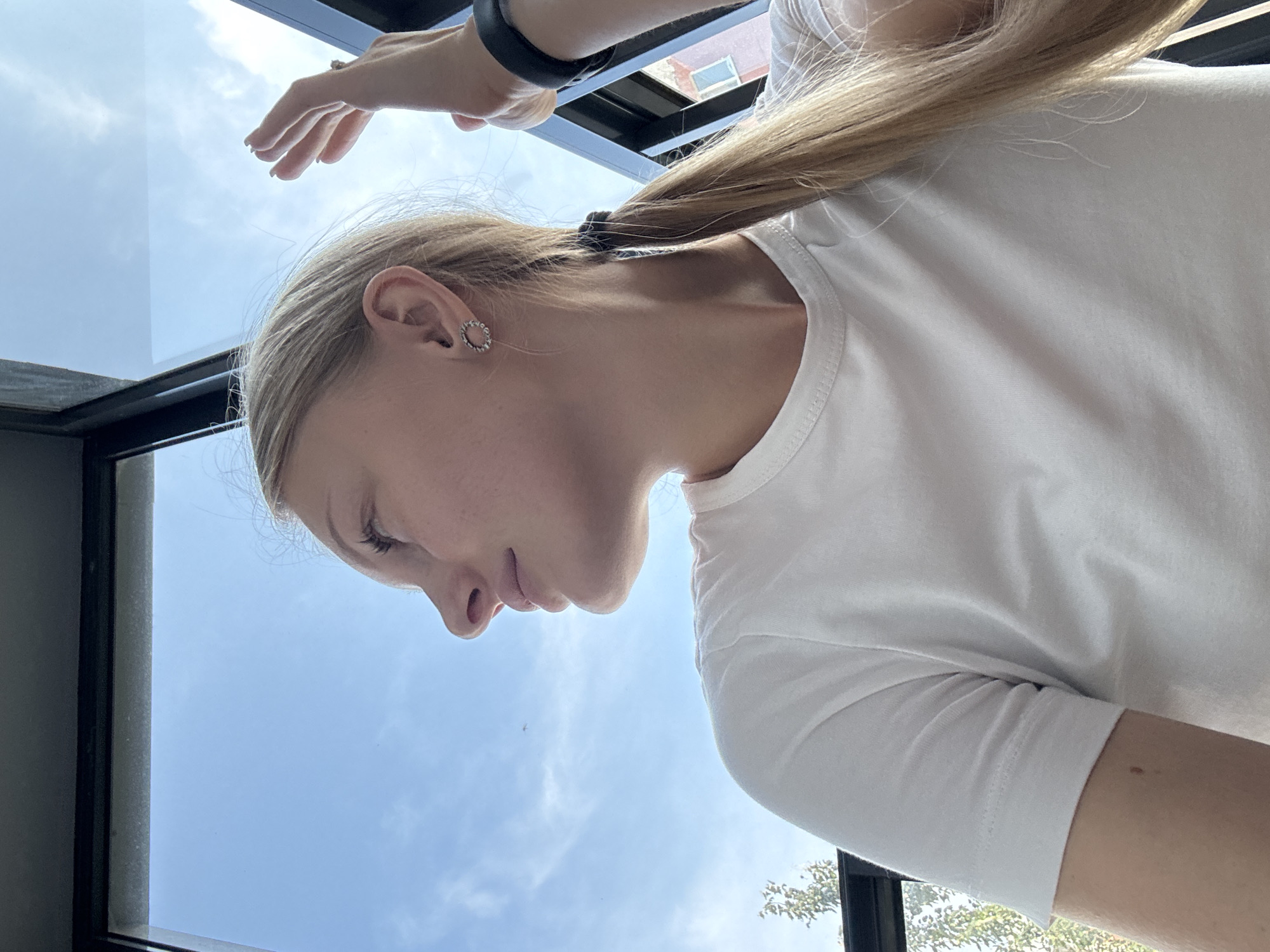 A young woman with blonde hair pulled back in a ponytail is shown from the shoulders up, looking out a window. She is wearing a white t-shirt and a circular earring in her left ear. Her right hand is raised, with fingers splayed, partially obscuring a black fitness tracker on her wrist. The background is a bright blue sky with some wispy clouds, and a glimpse of buildings and trees can be seen outside the window. The scene appears to be set in a modern building with dark-framed windows, likely in an urban environment. While the specific location in Perm, Russia, cannot be determined from the image alone, the presence of outdoor elements suggests an accessible vantage point within the city.