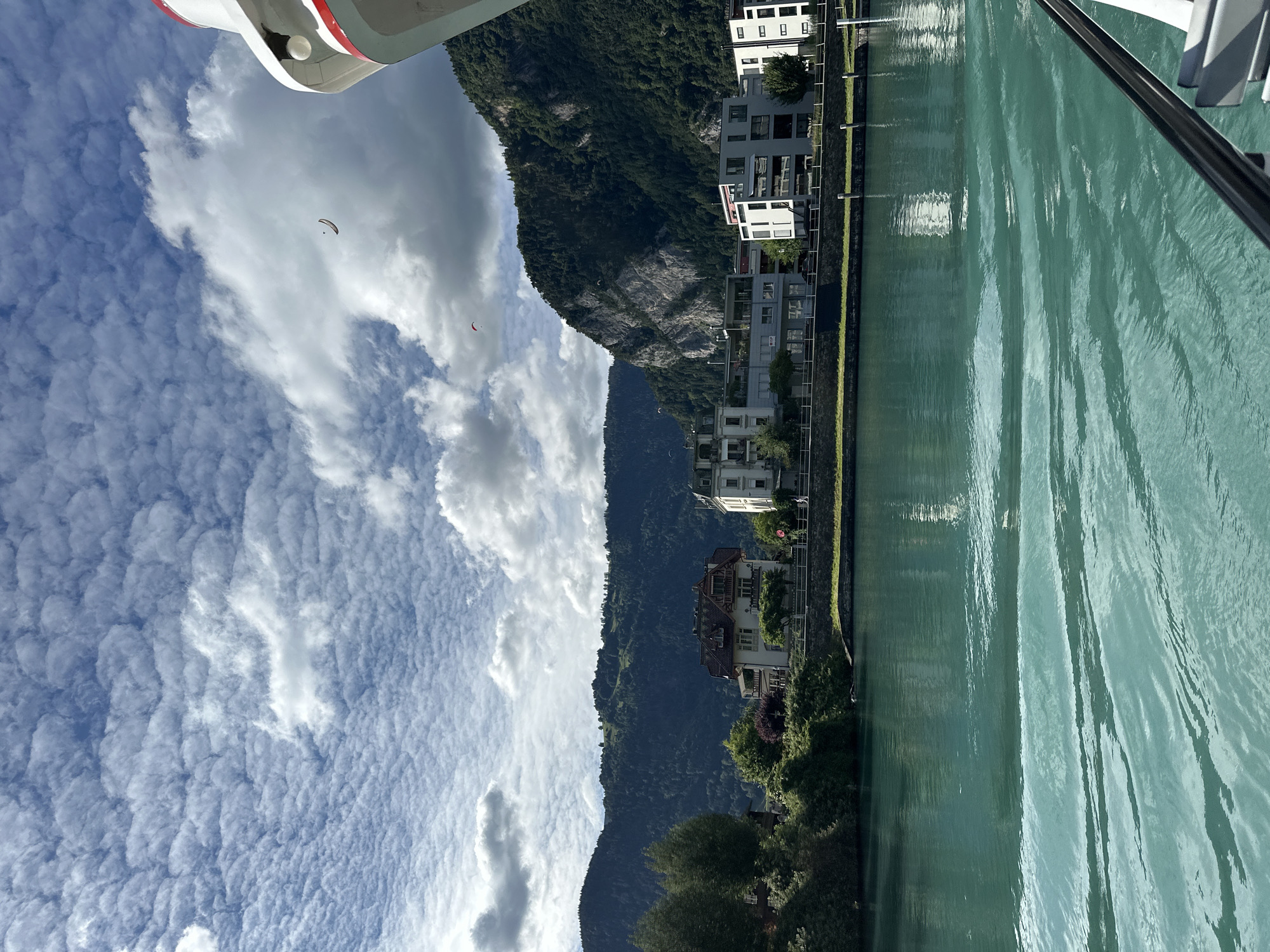This image depicts a waterfront scene in Interlaken, Switzerland, likely from a boat. In the foreground, turquoise water with gentle ripples fills the lower half of the frame, with the edge of a vessel visible at the bottom right. Across the water, a town or city area is visible, featuring a row of buildings with modern and traditional architecture. Behind the buildings, a lush, tree-covered mountain rises steeply, with rocky outcrops visible on its slopes. The sky above is a vibrant blue, filled with a mix of fluffy white clouds and some scattered cirrus formations. Two paragliders are visible in the sky, suggesting recreational activity in the area. The overall atmosphere is serene and picturesque, characteristic of the Swiss Alps region.
