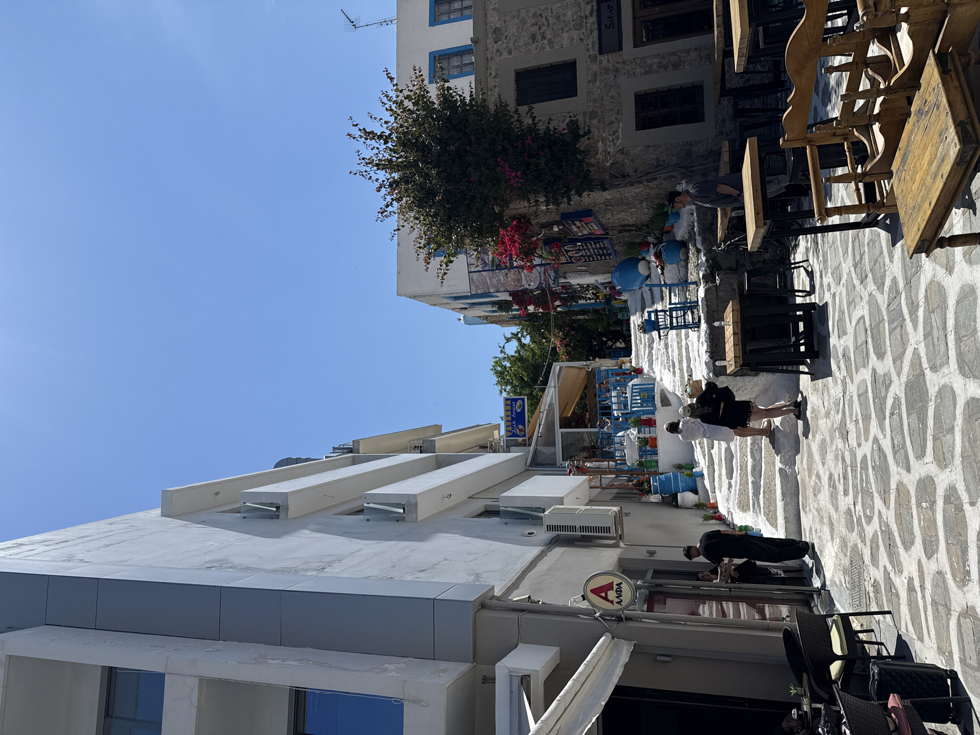 The image depicts a street scene in Kos, Greece, characterized by whitewashed cobblestone pathways. Along the street, people are walking. One individual is dressed in a black outfit. A pair of people are walking in white. Outdoor dining areas are visible, with wooden tables and chairs set up along the walkway. Buildings line the street, including a modern white structure with geometric roof protrusions, and more traditional buildings with stone facades and blue window frames. An "Alfa" sign is attached to the side of one building. Overhead is a clear blue sky, and vegetation, including a tree with red flowers, adds greenery to the urban landscape.