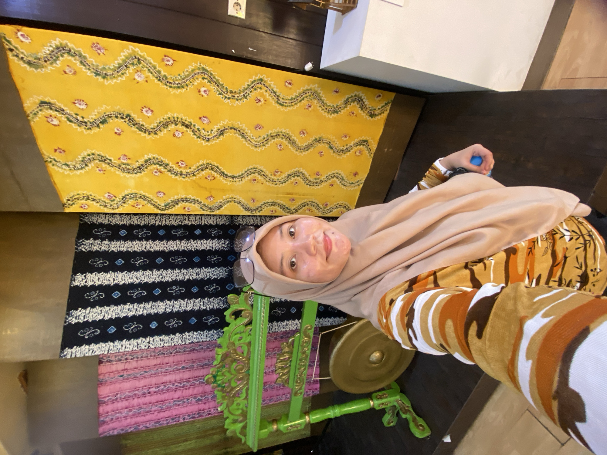 A woman wearing a light brown hijab and a camouflage-patterned shirt takes a selfie in front of traditional Indonesian textiles and a musical instrument. The textiles behind her are vibrantly colored: a green one on the far left, followed by pink, then black with white and blue diamond patterns, and a large yellow one with a repeating wavy pattern of green, pink, and purple motifs. To her left, a green and gold carved wooden stand holds a brass gong. The woman is smiling and has glasses perched on her head. She is holding a small blue object in her right hand. The background suggests an indoor cultural setting, possibly a museum or traditional house. This scene is located in Banjarmasin, Indonesia, a city known for its vibrant cultural heritage and traditional crafts.