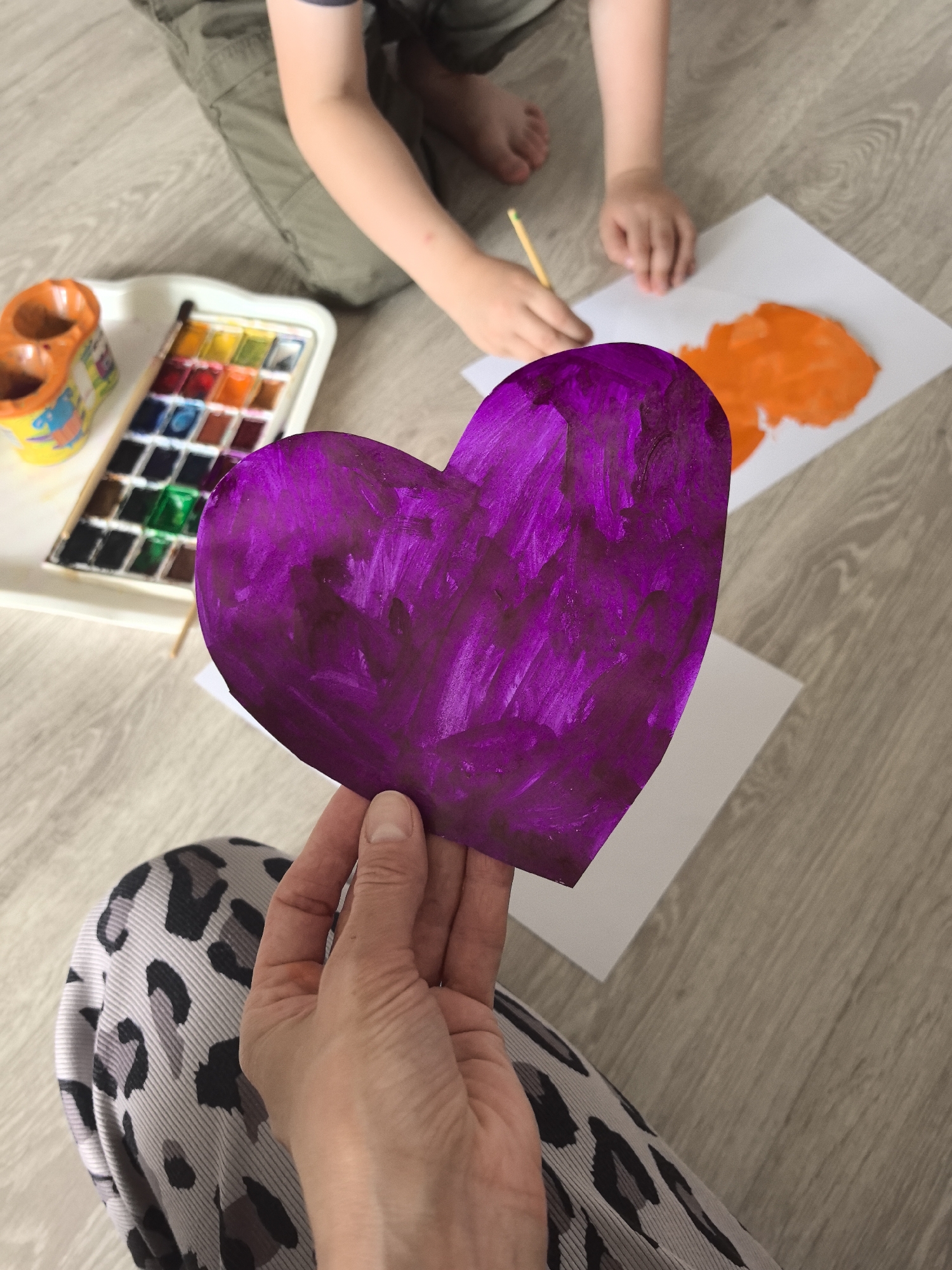 The image captures an indoor scene from a high-angle perspective, showing an adult and a child engaged in a creative activity on a light wooden floor, likely in a home in Perm, Russia.
In the foreground, an adult's hand holds up a heart-shaped piece of paper, vibrantly painted in a deep purple hue with visible brushstrokes, suggesting it's either a completed craft or a work in progress. The adult wears patterned pants, possibly leopard print, visible in the lower left.
In the background, a child is partially visible, kneeling or sitting on the floor. Their lower arms and hands are seen actively painting on a white sheet of paper using a paintbrush, creating an orange, irregular shape. The child is wearing green cargo-style pants.
To the left, a white plastic tray containing a watercolor paint set with multiple colorful pans is placed on the floor. Adjacent to the paint set is an orange, two-compartment container, likely used for water during painting.
The scene depicts a shared artistic moment, possibly a parent and child crafting together. The focus is on the act of painting and the creation of handmade items. There are no visible texts, and the time of day or external weather conditions cannot be determined from the indoor setting. The image conveys a sense of engagement and creative expression.