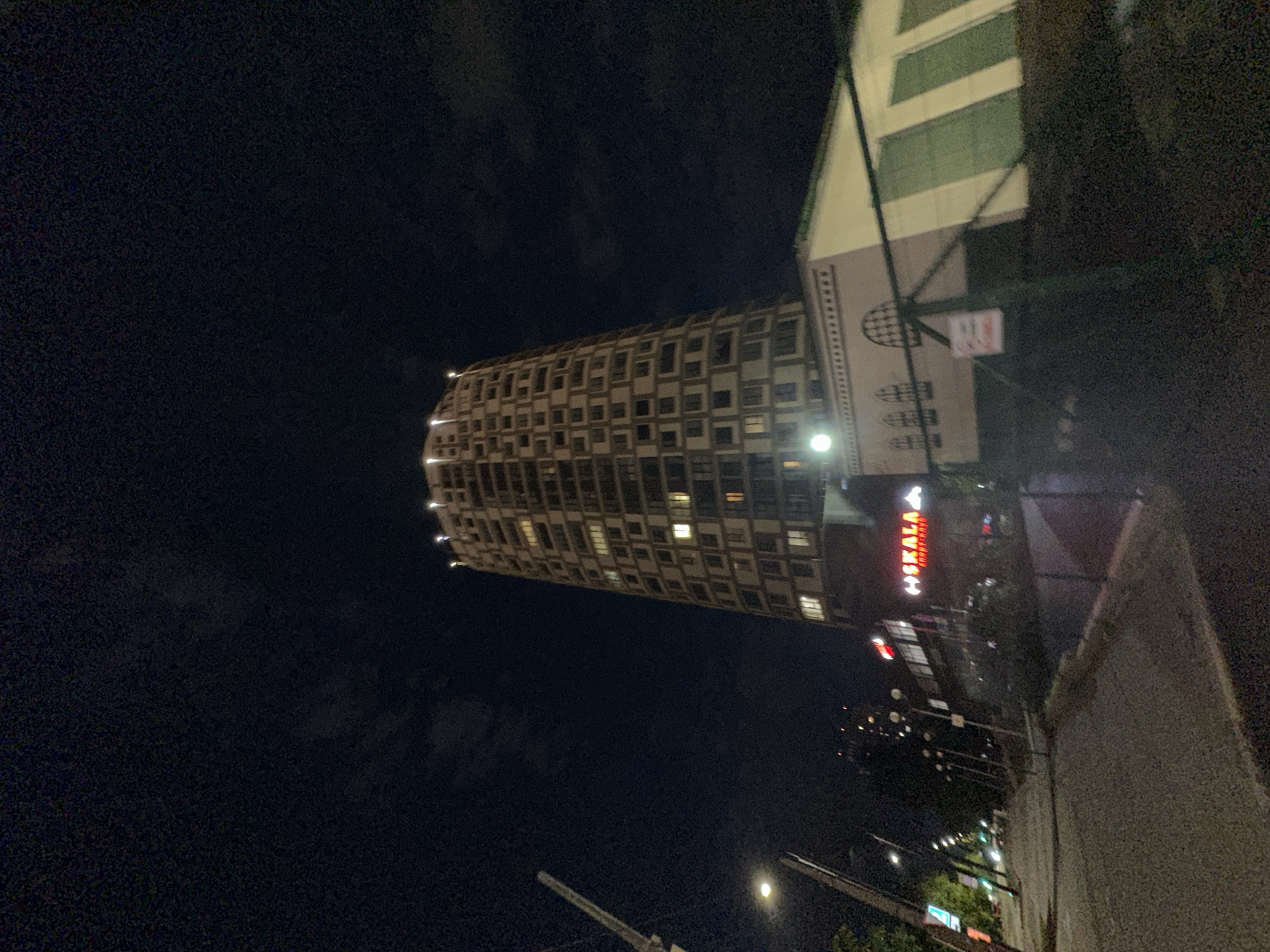 The image shows a night view in Perm, Russia. The distinct cylindrical building with numerous windows is the "Perm Telecom Building," a notable landmark. The "Skala" sports shop neon sign is visible in the middle of the image. The surroundings include a street with streetlights and some greenery. The sky is dark, suggesting it is nighttime. The shot is taken from street level, looking upwards at an angle.