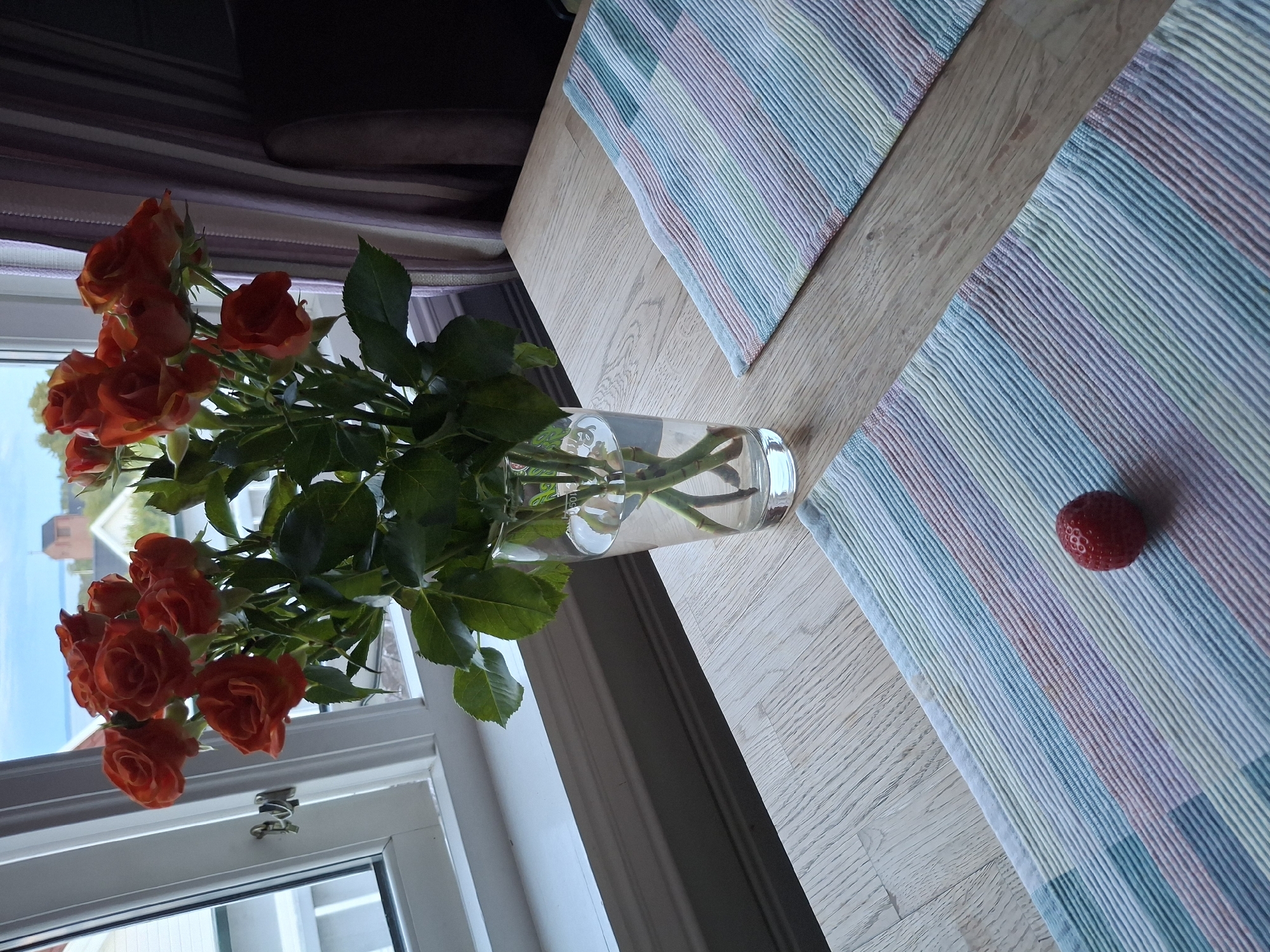 An indoor scene depicts a bouquet of orange roses in a clear glass vase filled with water, positioned on a light-colored wooden table. Two placemats with thin, multicolored stripes in shades of blue, green, pink, and yellow are laid out on the table. One strawberry rests on the lower placemat. In the background, a window reveals a view of buildings and the sky. The image is shot from a slightly elevated angle. The context provided places the scene in Åsgårdstrand, Norway.
