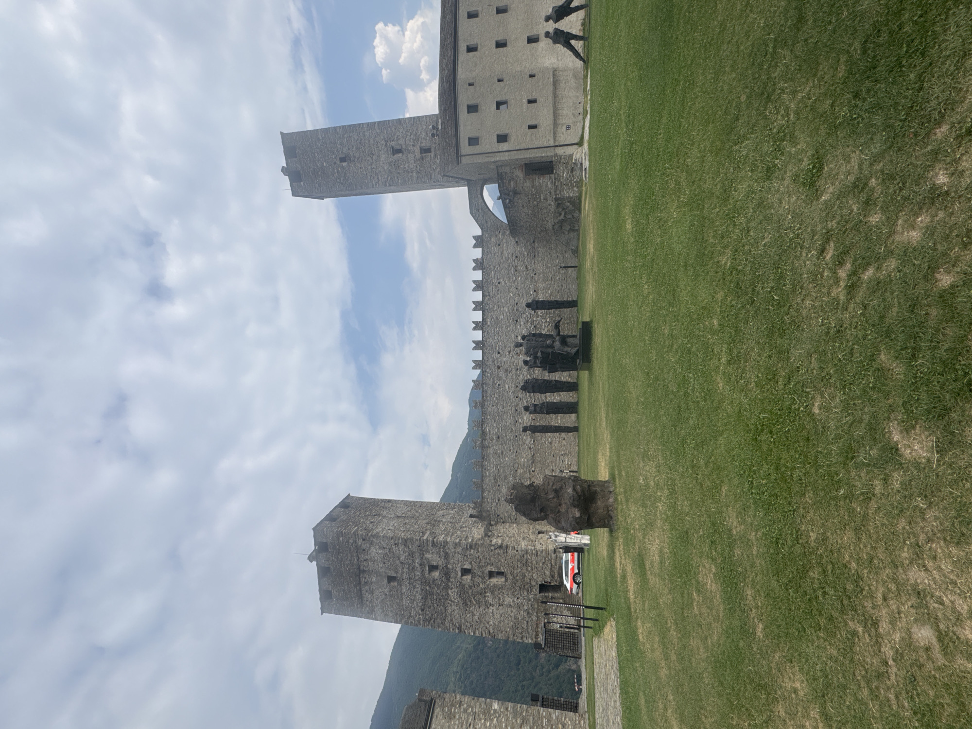 The image shows a view of Castel Grande in Bellinzona, Switzerland. In the foreground, there is a well-maintained grassy area with several abstract sculptures. In the middle ground, the stone walls and towers of the castle are visible, along with an arched passageway. A white and red vehicle, possibly an emergency vehicle, is parked near the entrance to the castle grounds. In the background, a green, forested mountain range rises against a partly cloudy sky. The overall scene depicts a historical landmark with artistic installations in a natural setting.