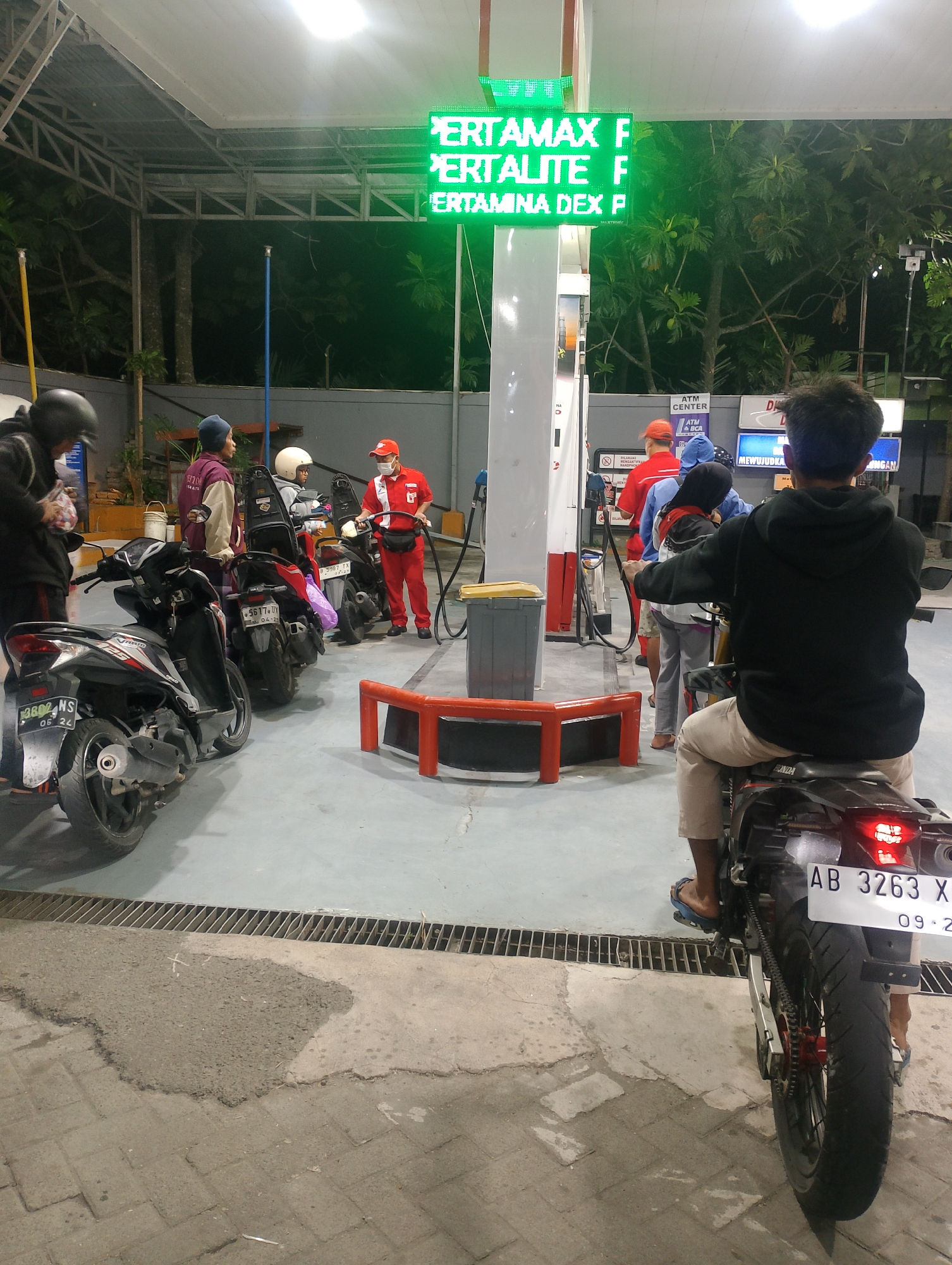 The image depicts a busy nighttime scene at a gas station in Harjobinangun, Indonesia. A green LED sign at the top of a gas pump displays fuel prices: "PERTAMAX F," "PERTALITE F," and "PERTAMINA DEX P." Several individuals are present, including a gas station attendant in a red uniform and face mask dispensing fuel to a person on a motorcycle. Another person on a motorcycle, wearing a black hoodie and tan pants, is facing away from the camera, while others are gathered near other motorcycles. The ground is paved with concrete and interlocking bricks. The environment appears to be an outdoor, covered area of the gas station, with some trees visible in the background.