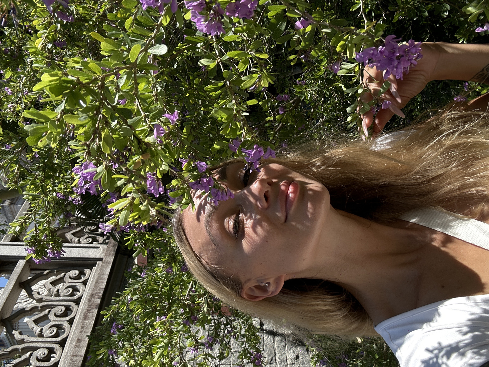 A woman is lying down under a flowering bush. The bush has green leaves and purple flowers. The woman has long blond hair. She is wearing a white shirt. There is a carved stone structure in the background. The location is likely outdoors, possibly in a garden or park in Dĩ An, Vietnam.