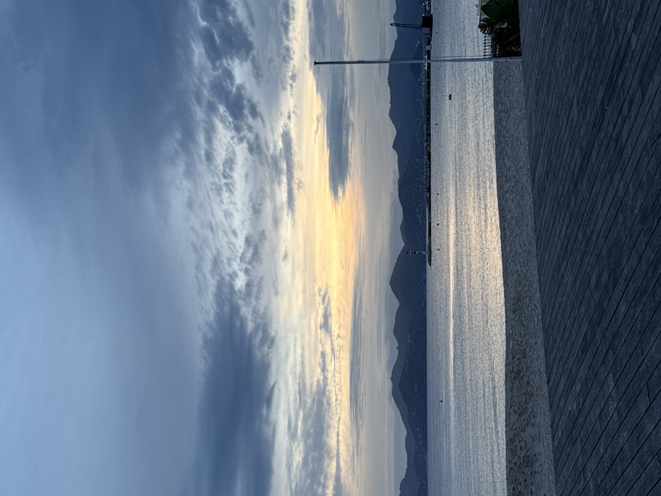 The image displays a serene coastal landscape, likely in Cannes, France, captured during either sunrise or sunset. The sky occupies the upper portion, featuring a mix of dramatic dark and lighter, wispy clouds. These clouds gradually transition into a soft, golden glow near the horizon on the right, indicating the sun's position.
The tranquil surface of a large body of water, presumably the Mediterranean Sea, stretches across the middle of the frame, beautifully reflecting the warm light from the sky. In the distance, dark, silhouetted mountains or hills line the far shore, appearing as a soft blue-grey against the luminous horizon. Along this distant coastline, faint lights or structures are visible, including a small white lighthouse or navigational beacon. Towards the far left, a distinct harbor crane suggests the presence of a port.
In the mid-ground, a tall, slender pole, possibly a light fixture or mast, rises vertically from the water's edge. The immediate foreground on the right features a broad, textured surface, appearing to be a dark wooden or tiled promenade or pier, running parallel to the water. A hint of green foliage, resembling palm leaves, is visible behind this foreground structure. The scene is devoid of explicit human activity, conveying a peaceful and calm atmosphere. No discernible text is present in the image.