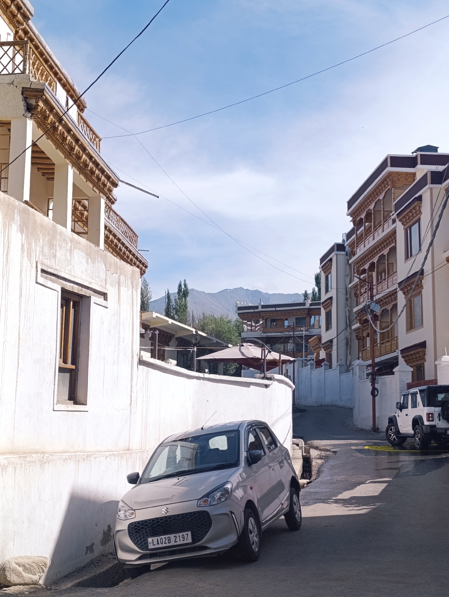 The image depicts a street scene in Leh, India, characterized by traditional Ladakhi architecture. In the foreground, a silver Maruti Suzuki Celerio car is parked diagonally on the left side of the narrow street, facing towards the right. Its license plate reads "LA02B 2197". Further down the street on the right, a white Jeep Wrangler is parked. The buildings on both sides of the street feature multi-story structures with ornate wooden balconies and windows, typical of the region. The left side of the street is dominated by a white building with a balcony and a window. The background reveals distant mountains under a clear blue sky with scattered clouds. Power lines are visible crisscrossing the sky. The street surface appears to be asphalt, with some patches of water or dampness.