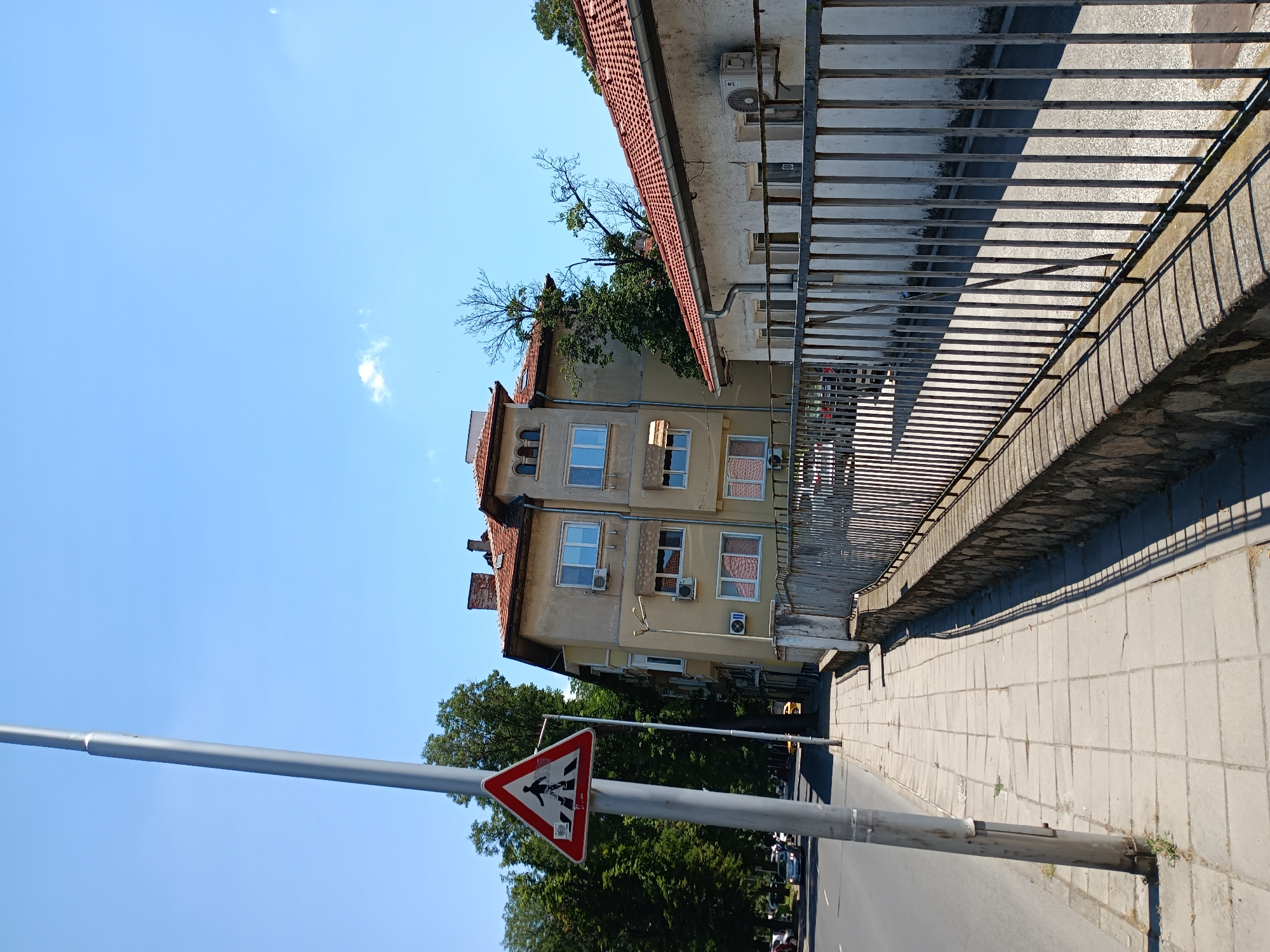The image shows a street scene in Plovdiv, Bulgaria. The photograph is taken from a low angle, looking up at a multi-story building with a red-tiled roof. The building is painted a light yellow and has several windows, some of which appear to be air-conditioned units. A metal railing runs along the side of the building, casting a shadow on the paved sidewalk below.
In the foreground, a street sign with a red border and a white triangle depicts a pedestrian crossing symbol, indicating a designated area for people to cross the street. A tall metal pole supports this sign, extending out of the frame. The sky is a clear blue with a few wispy clouds. Trees with lush green foliage are visible behind the building, and a street with parked cars is to the left. The sunlight suggests it is daytime, likely midday or early afternoon, given the strong shadows. The scene appears peaceful and ordinary, capturing a moment of everyday urban life. There is no visible text other than the symbols on the street sign.