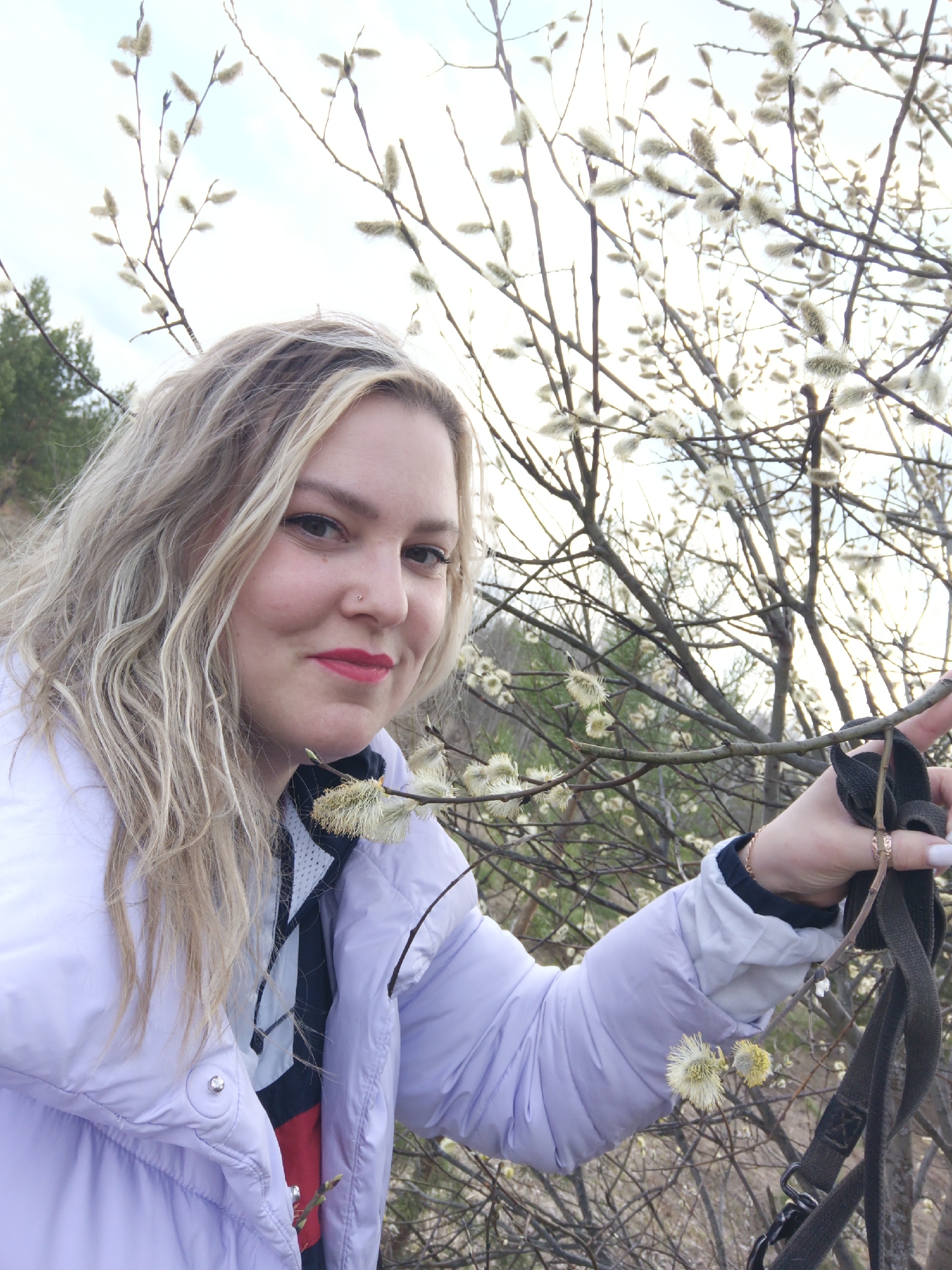 The image is a selfie taken in a location with pussy willow trees. A woman with blonde hair, red lipstick, and a nose piercing is the main subject. She is wearing a light purple puffer jacket and holding a black strap. The background consists of a cloudy sky, trees, and branches with pussy willows. The location is in the city of Марковский, Russia.