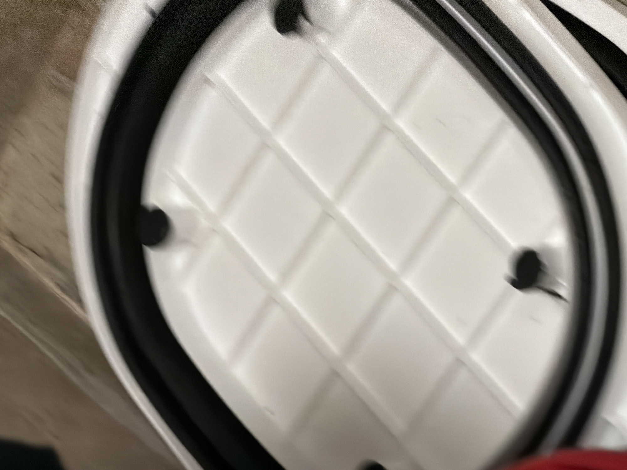 The image shows a close-up, blurry view of a white plastic lid with a grid pattern on its surface, bordered by a black rubber seal. There are several black circular protrusions on the white surface, possibly mounting points or fasteners. The lid appears to be part of a larger object, as suggested by the layered white and black plastic structure visible at the bottom right. The background is a soft, out-of-focus brown texture, possibly a carpet or fabric. There are no people or specific landmarks visible to indicate a location in Sofia, Bulgaria. The image is an indoor, close-up shot of an object.