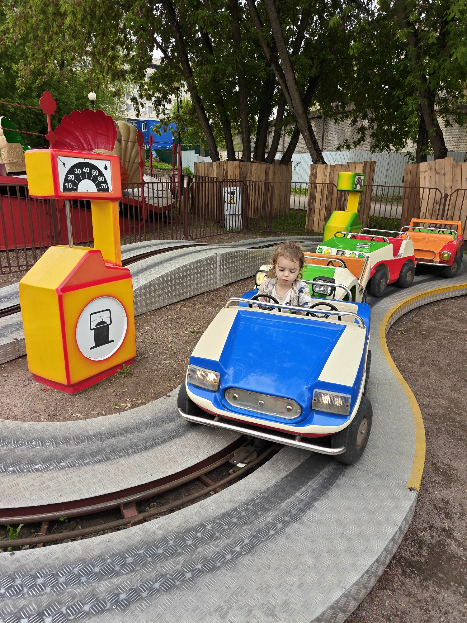A young girl is operating a blue and cream-colored children's car ride on a circular track in what appears to be a park or playground. The car is in the foreground, followed by green, red and white, and orange cars connected in a train-like fashion. A mock-up gas station painted in yellow and red stands beside the track, including a meter and a pump. The track consists of a diamond-plated surface and a metal rail. The setting is outdoors, with trees and buildings visible in the background. The location is Moscow, Russia.
