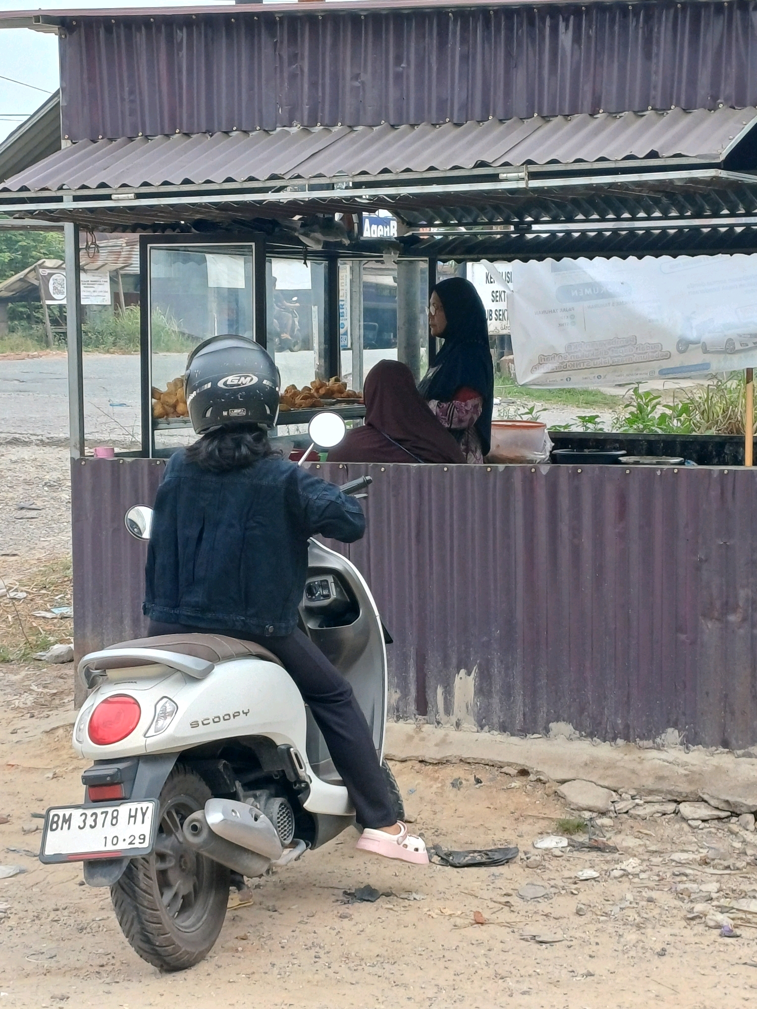 In Dumai City, Indonesia, a person wearing a dark blue denim jacket and a black helmet is seated on a white Honda Scoopy scooter, facing away from the camera. The scooter's license plate reads "BM 3378 HY 10-29". In the background, a food stall with a corrugated iron roof is visible. A person wearing a dark headscarf and a brown head covering is standing behind the counter of the food stall, which displays fried food items in a glass case. The area around the stall is unpaved, with dirt and scattered debris.