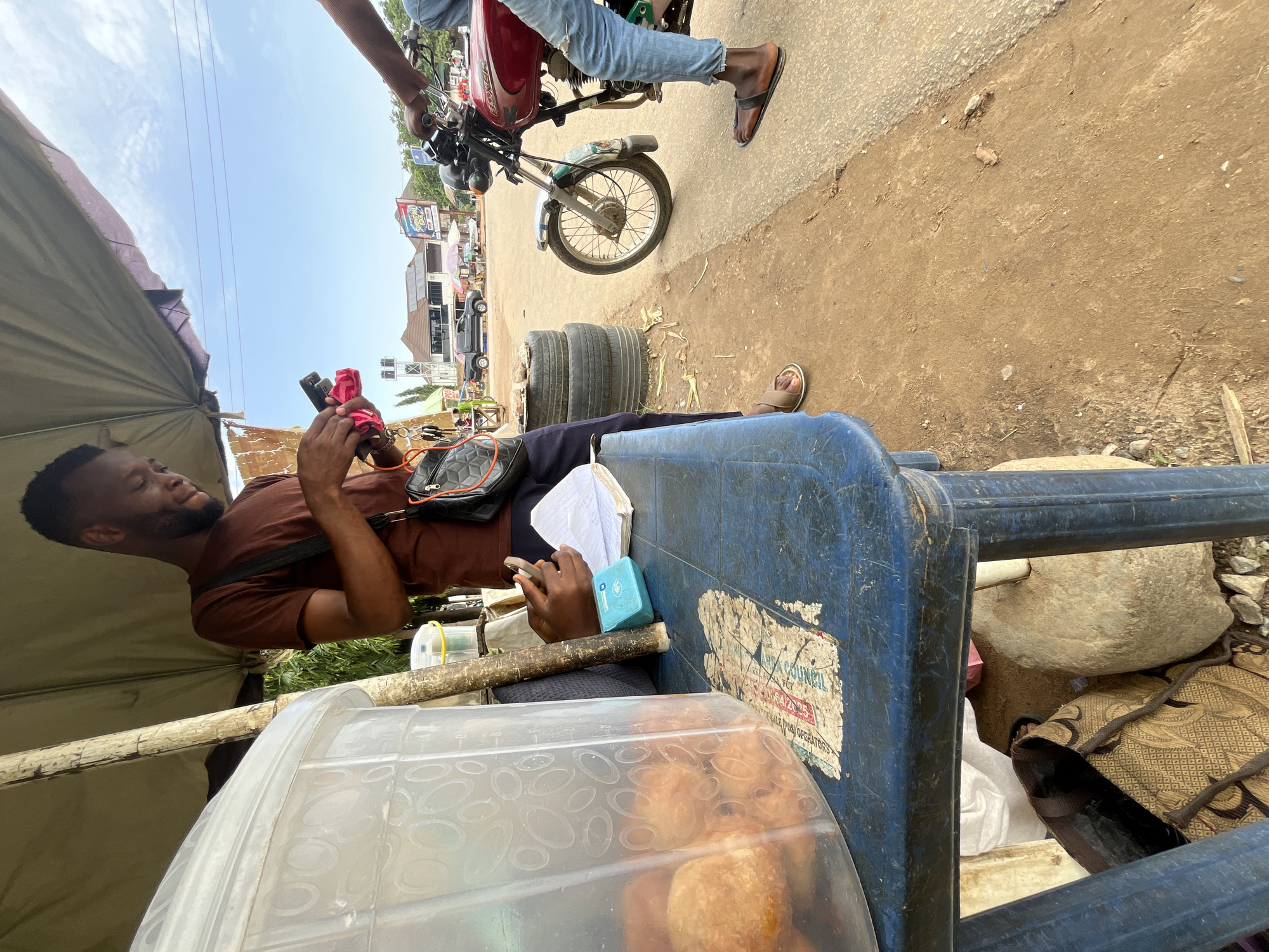 In Abuja, Nigeria, a man in a brown t-shirt stands under a canopy, holding a smartphone and a pink object, possibly a cloth or another phone. He has a black shoulder bag slung across his body. To his left, on a blue table, is a clear plastic container filled with fried snacks, a small blue packet, and an open notebook. A second person, partially visible on the right, is seated on a red motorcycle and wearing blue jeans. The background shows a street scene with buildings, other vehicles, and a clear sky. The ground is a mix of dirt and asphalt.
