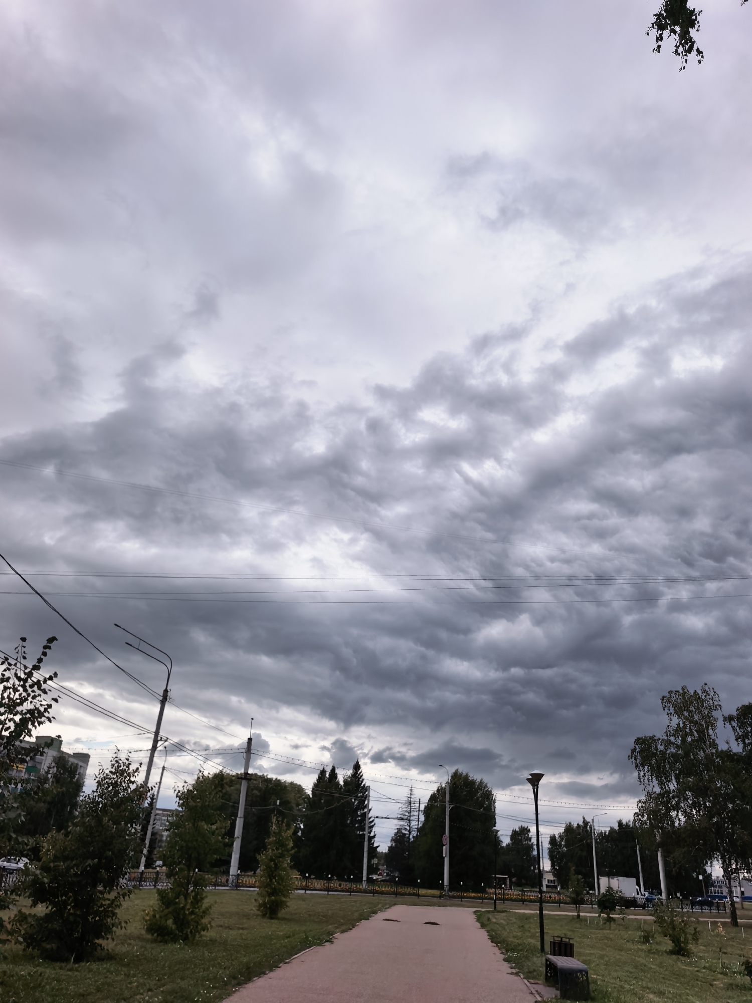 This image depicts a cloudy sky over a park area in Sterlitamak, Russia. The sky is filled with heavy, grey clouds, suggesting an overcast or stormy day. Below the sky, there are overhead power lines and utility poles. The park features a paved walkway, grassy areas, and several trees, some of which are mature and others are saplings. In the background, apartment buildings and more trees are visible. The overall scene is one of a somewhat gloomy, urban park setting.