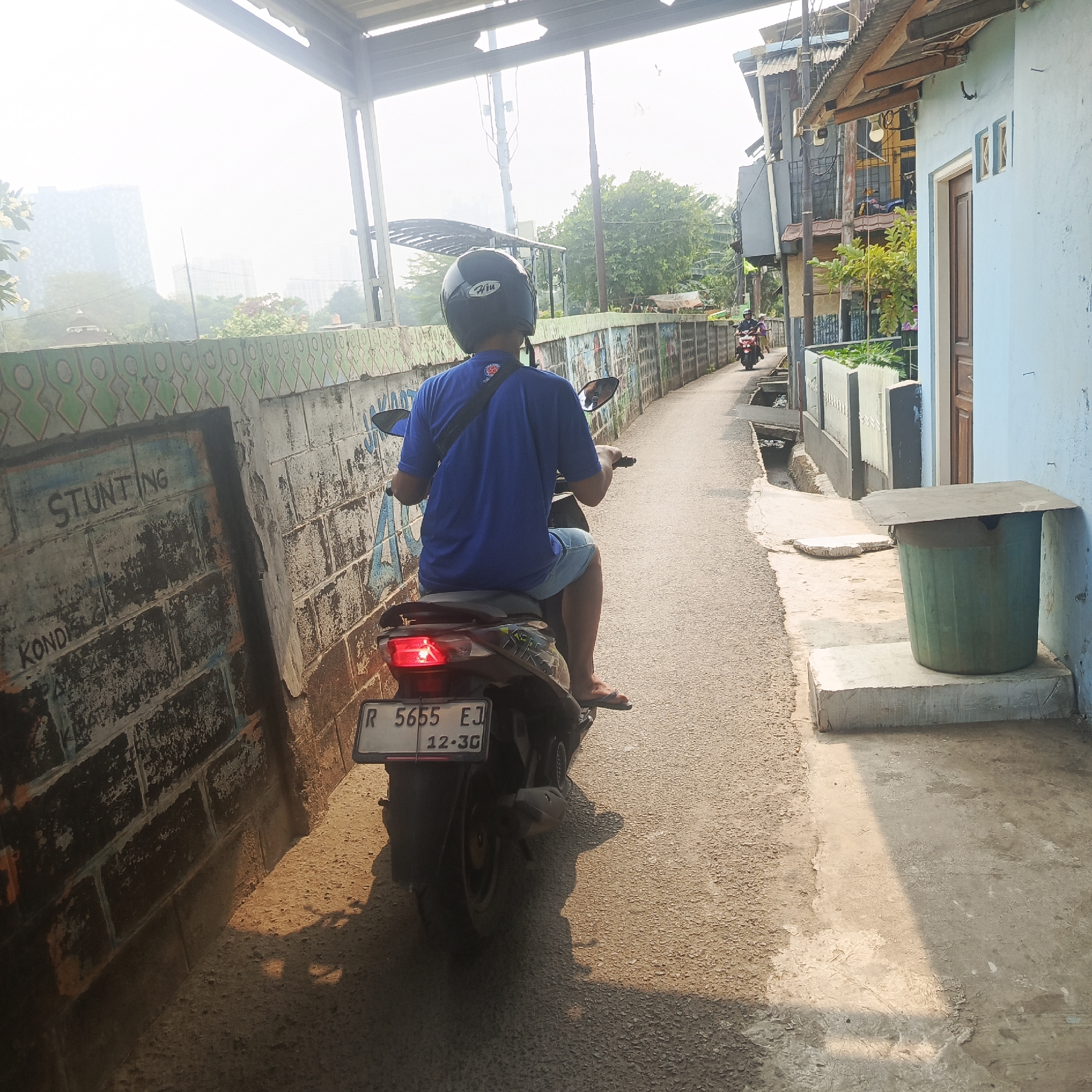 A person wearing a blue shirt and shorts rides a scooter down a narrow paved alleyway. The person has a black helmet on and is seen from behind. The alley is bordered by a low wall covered in graffiti on the left and a light blue building with a brown door on the right. In the background, other buildings and trees are visible. The sky is bright and hazy. A license plate on the scooter reads "R 5655 EJ 12-30". The scene appears to be in an urban environment, possibly in the Menteng Dalam area of South Jakarta, Indonesia, characterized by densely packed housing and narrow access roads.