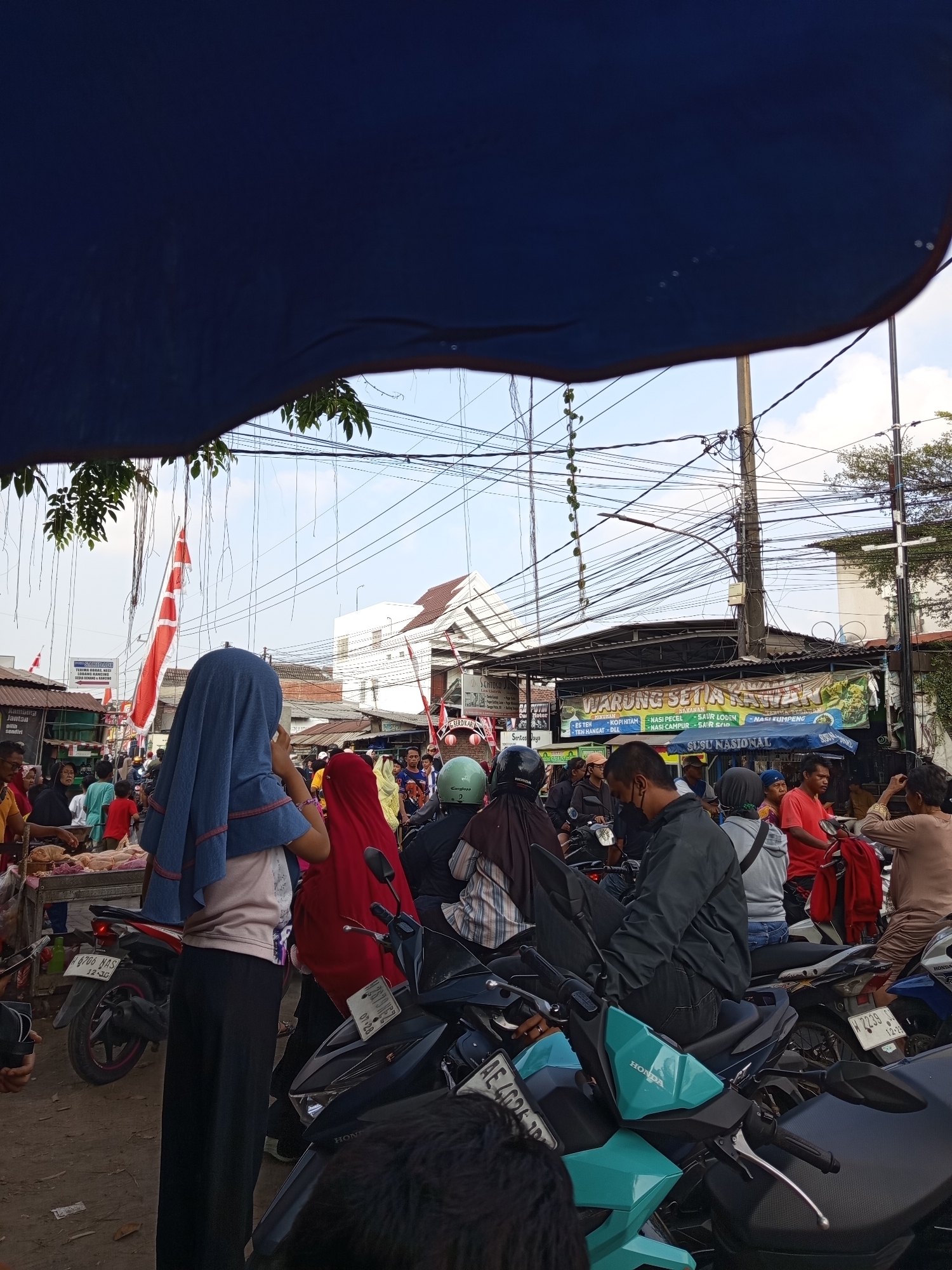 The image depicts a crowded street scene in Suko, Indonesia. The foreground is dominated by the dark blue canopy of an overhead structure. In the mid-ground, a group of people are gathered, many on motorcycles. A woman in a light blue hijab is looking to her right, while a woman in a red hijab stands nearby. Several motorcycles are parked and being ridden, indicating a bustling environment. A stall selling raw meat is visible on the left, with people examining the produce. Behind the people and motorcycles, buildings and electrical wires are prominent. A banner reading "WARUNG SETIA KAWAN" is displayed on a building, suggesting a food establishment. Red and white flags are also visible, possibly indicating a local event or celebration. The ground is paved with dirt and gravel. The sky is partly cloudy.