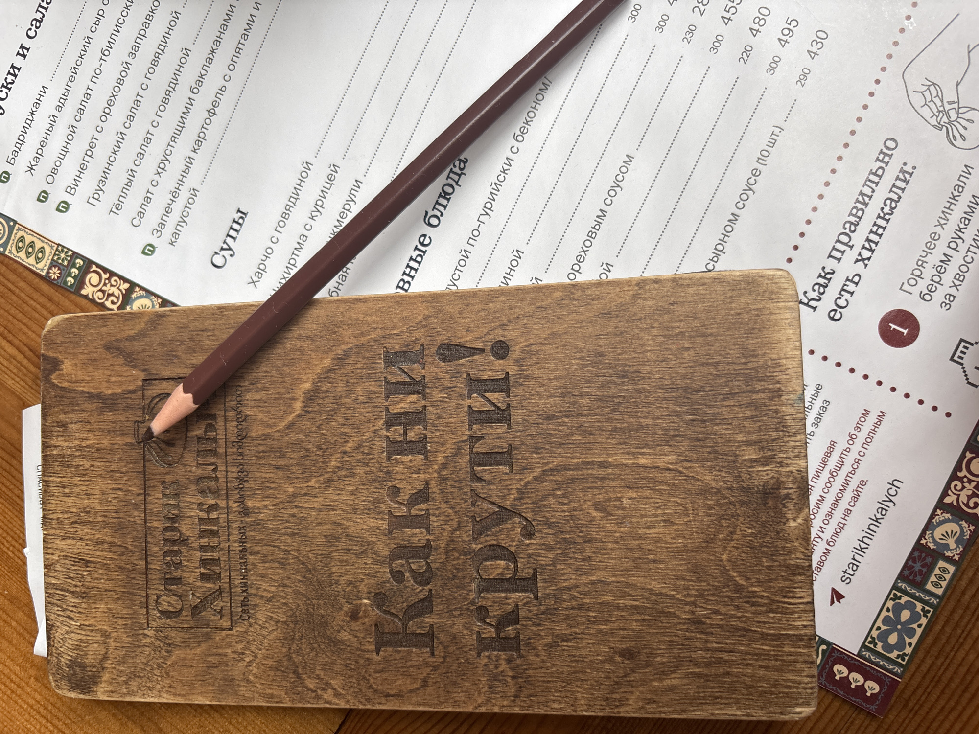 Here is a description of the image:
The image depicts a menu from the restaurant "Starik Hinkalych" in Izhevsk, Russia. The menu has a wooden cover with the name of the restaurant engraved in Russian, alongside the phrase "Как ни крути!" (Kak ni kruti!), which roughly translates to "No matter how you twist it!".
On top of the menu rests a brown pencil, pointed towards the restaurant's logo. The open menu reveals the dishes offered, including soups like kharcho and various salads. The right side of the menu includes instructions on how to properly eat khinkali (Georgian dumplings). The image suggests a cozy and possibly traditional Georgian dining experience.