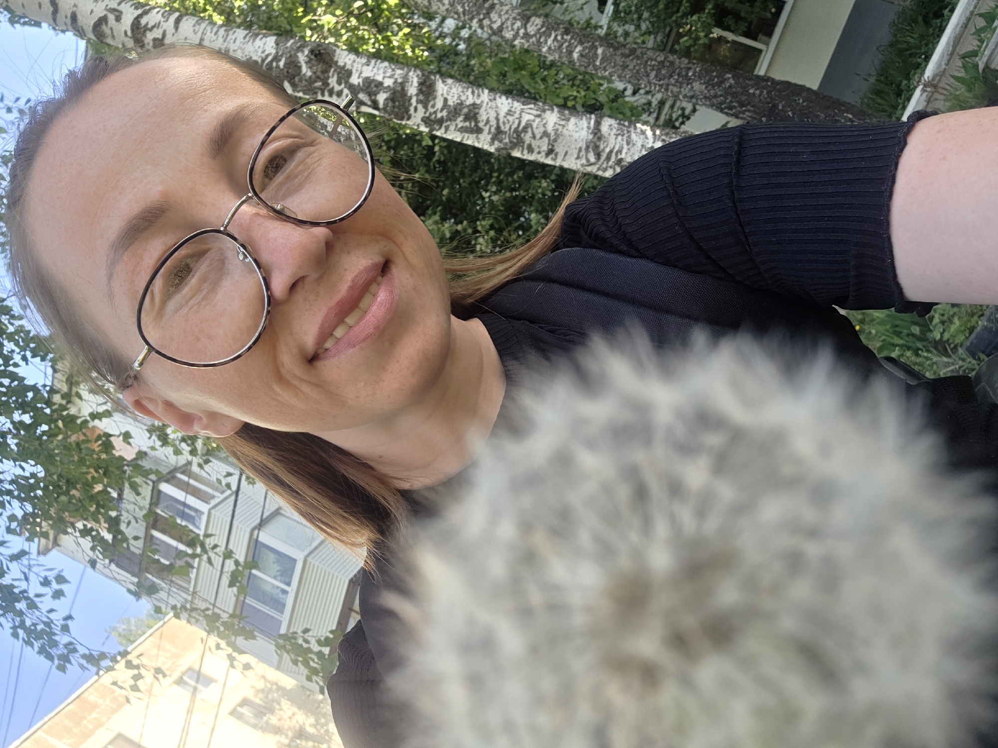 A woman with glasses is taking a selfie outdoors in Chaikovsky, Russia. The woman has light brown hair and is wearing a black top. She is smiling and holding a large, fluffy dandelion seed head in front of the camera, obscuring part of the view. Behind her, there are birch trees with distinctive white bark. In the background, buildings and a blue sky can be seen. The woman's face is well-lit, and the dandelion is out of focus due to its proximity to the camera.