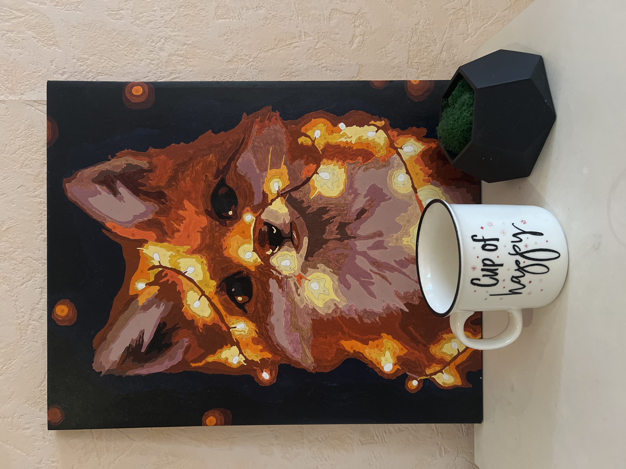 A painting of a fox wearing Christmas lights hangs on a textured beige wall. The fox is depicted in shades of brown, orange, and tan against a dark blue/black background. The lights are painted in yellow and white, scattered across the fox's face and body. Dark brown circles surround the painting. A black, geometric wall planter containing green moss is placed above the painting. A white mug with the text "Cup of happy" in black script is positioned to the right of the painting and below the wall planter. There is no indication of the image being taken in Votkinsk, Russia.