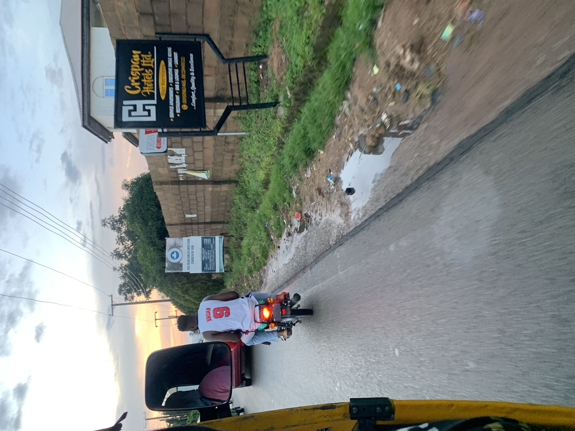 The image captures a street scene in Gura Top, Nigeria, likely during the late afternoon given the soft, diffused light and the presence of clouds. The perspective is from within a vehicle, possibly a tricycle, as indicated by the yellow painted frame on the left side of the image. In the foreground, the paved road shows signs of moisture, with several puddles reflecting the overcast sky. The roadside is lined with patchy green grass and scattered debris, including plastic bags and other refuse.
Further down the road, a man wearing a white jersey with "JORDAN" and the number "9" emblazoned on it is riding a motorcycle. He is wearing blue jeans and brown shoes. He is accompanied by another person sitting behind him, partially obscured, wearing a maroon garment. The motorcycle's brake light is illuminated, indicating it might be slowing down or stopped.
On the right side of the road, a brick wall serves as a backdrop for two signs. The larger sign, black with yellow text, advertises "Crispan Hotels Ltd." and lists its services, including "Service Apartments," "Executive Single Rooms," "Restaurant," "Bar & Lounge," and "Laundry." Below this, it states "...Comfort, Quality & Excellence." A smaller sign, red and white, reads "JORDAN TOURIST LODGE." A building with a white facade and a light blue roof is visible behind the Crispan Hotels sign.
To the left of the Crispan Hotels sign, another sign is mounted on a brick wall, advertising "THE REDEEMED CHRISTIAN CHURCH OF GOD." This sign details church activities and service times. In the distance, trees and utility poles are visible against a pale sky streaked with hues of orange and pink from the setting sun. The overall environment suggests a somewhat developed urban or semi-urban area with a mixture of commercial establishments and residential buildings, alongside common issues of street cleanliness.