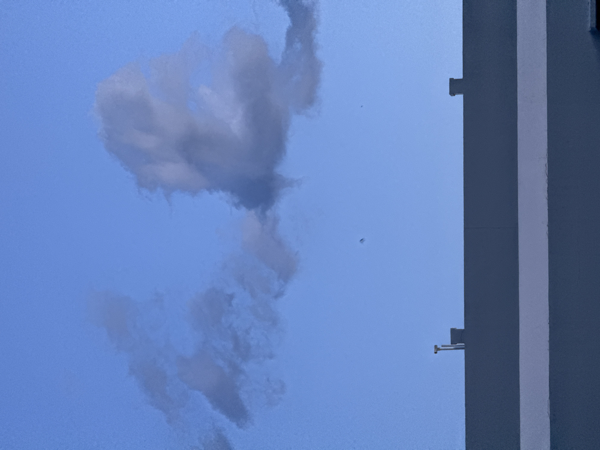 The image, taken from a low angle, shows a blue sky with a cluster of grayish-white clouds. The clouds form a vertical shape with a denser mass at the top and thinner tendrils extending downwards. A dark speck, possibly a bird or insect, is visible in the sky. A section of a building occupies the right side of the frame, featuring gray vertical structural elements. Protrusions extend from the building's surface, suggesting architectural details. The scene is consistent with an outdoor setting on a day with partial cloud cover. The location provided suggests a possible context, but the image does not provide concrete evidence to verify it.