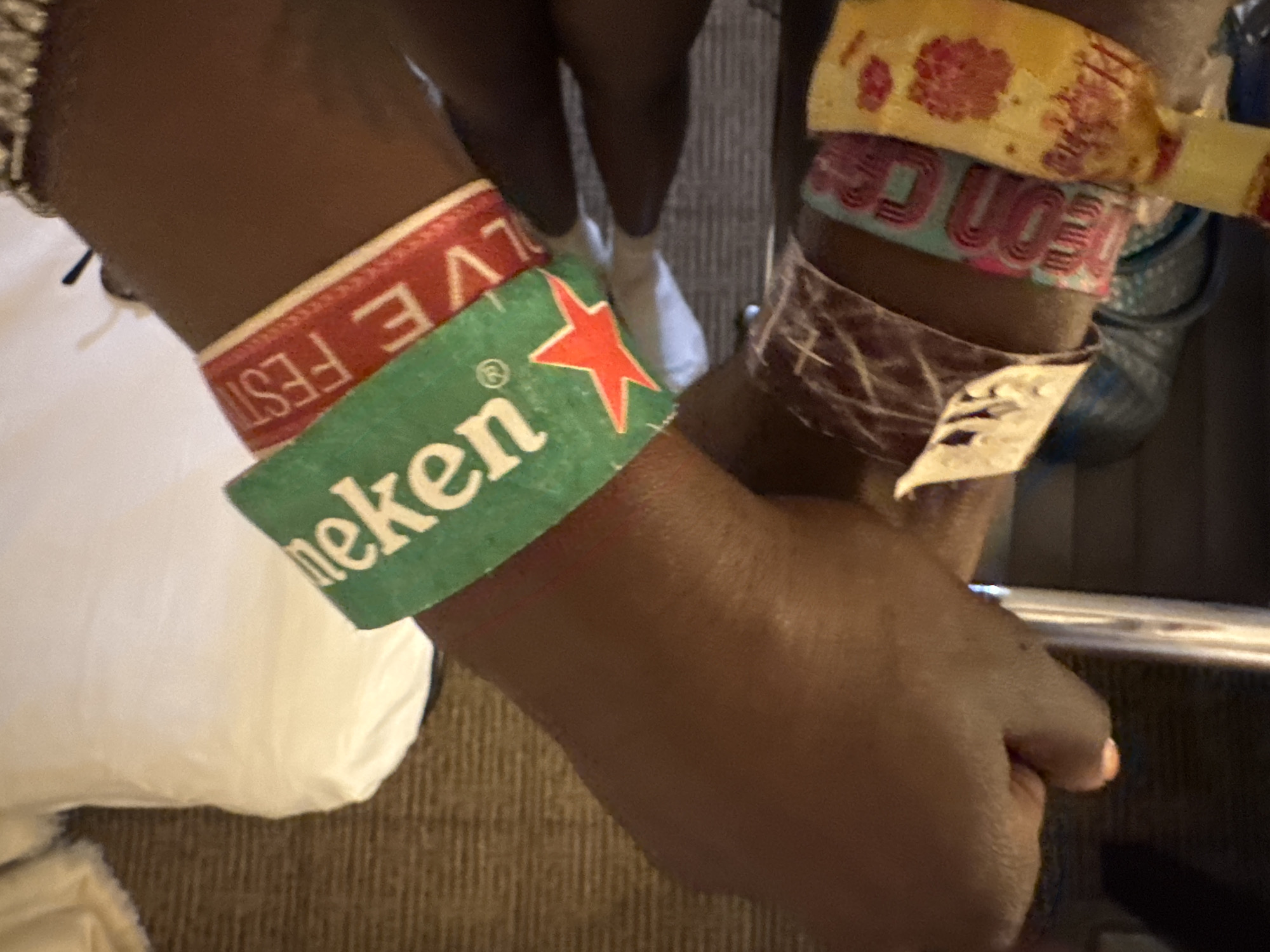 A close-up shot shows two dark-skinned arms with multiple wristbands on each, suggesting a recent attendance at various events. The left arm has a green wristband with the word "Heineken" and a red star visible, along with a red wristband labeled "LIVE FEST." The right arm sports multiple wristbands of different colors and designs, one appearing to read "MOTION." The scene is indoors, as indicated by the presence of a bed with a white sheet to the left and what seems to be a table or fixture with a metal edge in the background. The city of Palm Desert or the United States of America cannot be confirmed based solely on the given image.