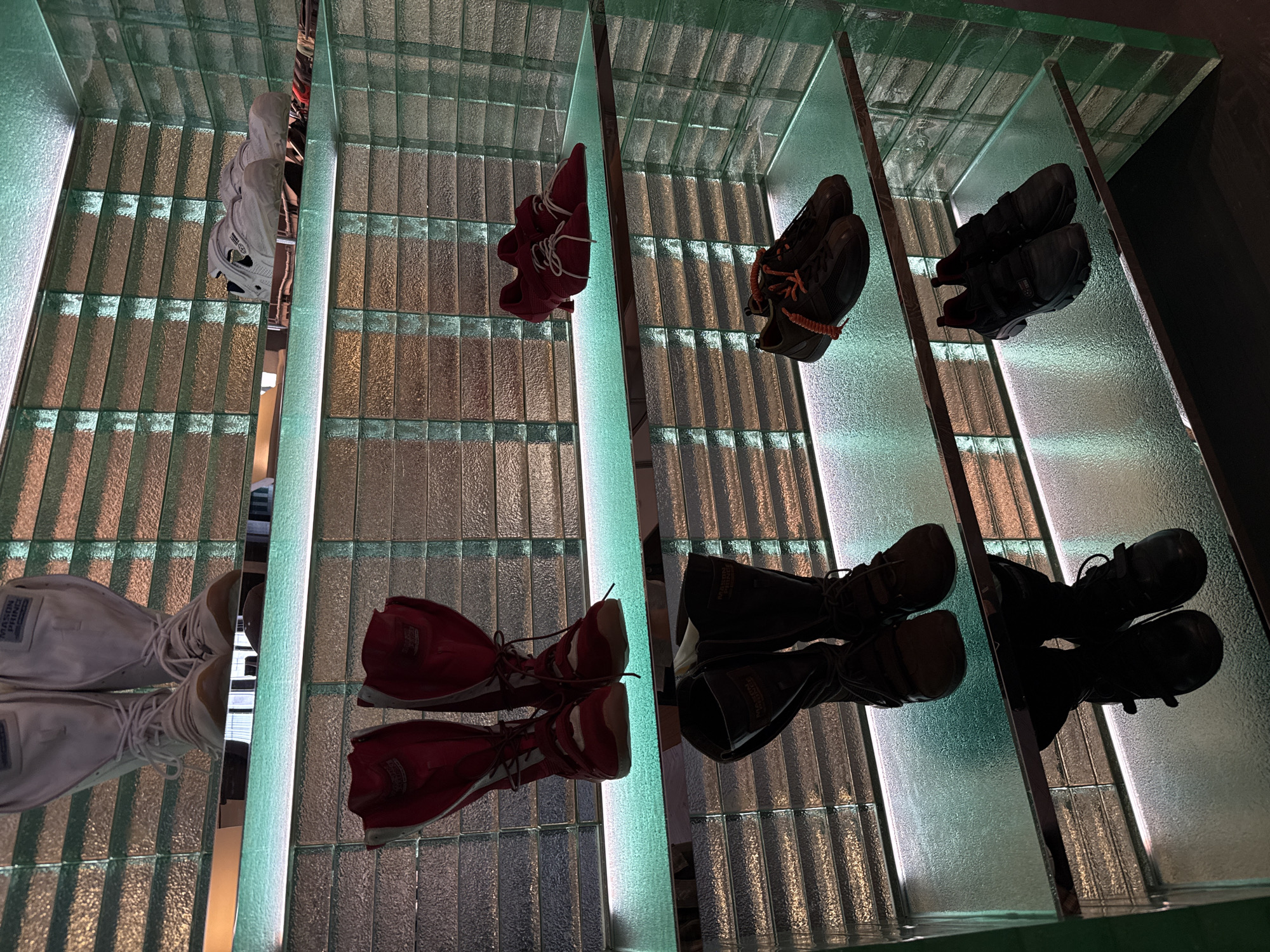The image shows a display of shoes hanging from shelves with a glass block background. There are four rows of shoes, each containing two pairs. The first row shows white shoes, the second shows red shoes, the third shows brown shoes, and the fourth shows black shoes. The shoes and the shelves are reflected on a glass surface. The location of this image is the city of Liwan District in China.