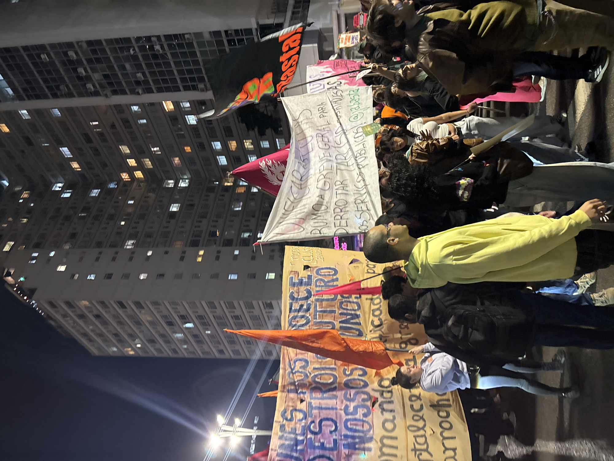 The image captures a night-time protest in what appears to be the city of São Paulo, Brazil, given the Portuguese language banners. The scene is dominated by a large group of people holding banners and flags. One banner is light in color with hand-written Portuguese text, calling for a general strike and demanding the defeat of public service tasks. Another banner, orange-toned, reads "DESTRO OUTRO NOSSOS UNOS," with additional text "mando cade po talece gre arroch." An orange pennant is also present. Individuals are wearing various clothing items, including a yellow hoodie and black jackets. In the background, a large high-rise building lit with numerous windows punctuates the night skyline, while streetlights cast a bright glare into the scene. The protest seems to be moving along a street or avenue.