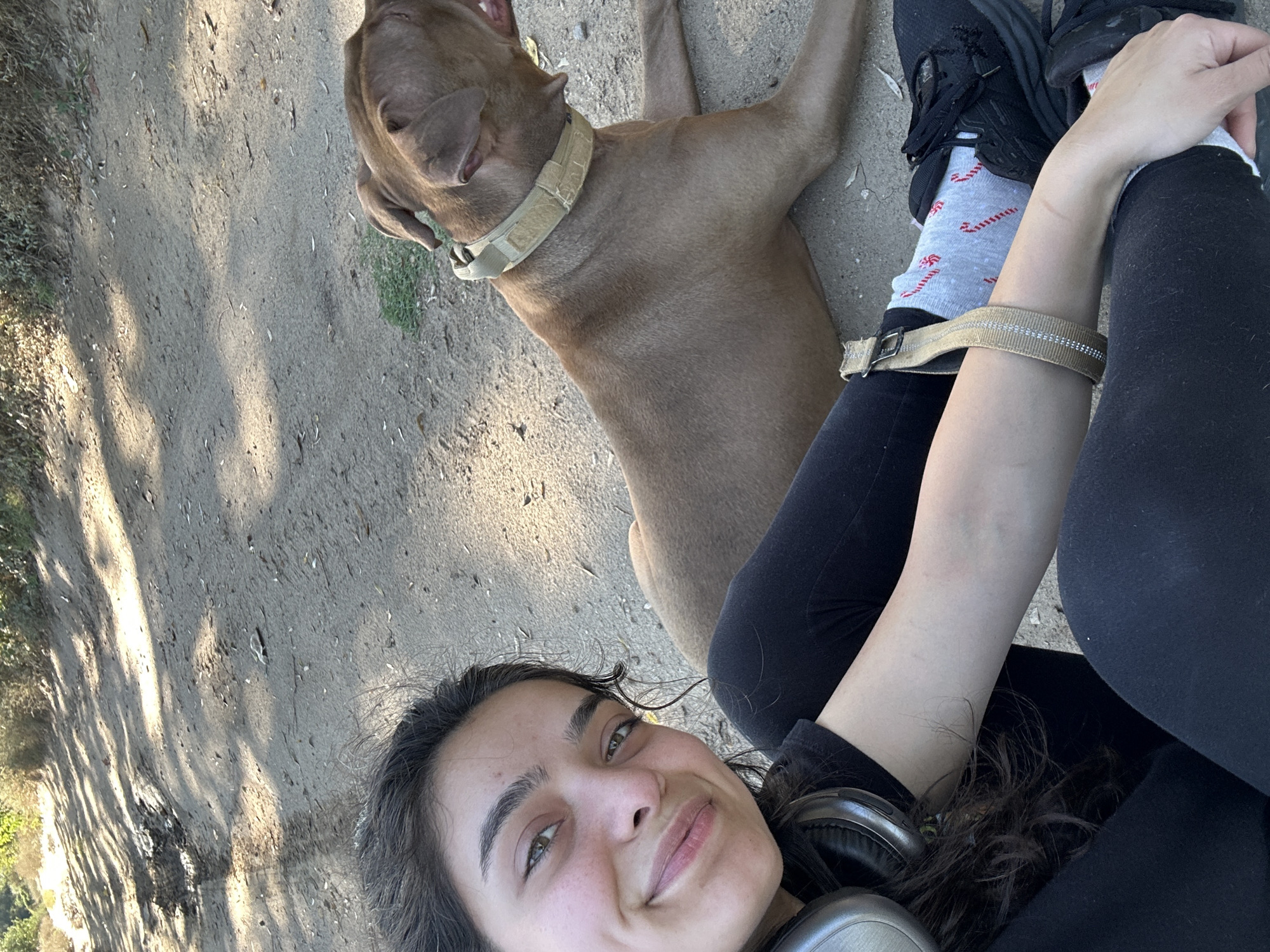In Malevo, Bulgaria, a young woman with dark, curly hair and light eyes smiles at the camera while sitting on a dirt path. She is wearing black athletic leggings, a black t-shirt, and headphones around her neck. Beside her, a brown pitbull lies on the ground. The dog wears a tan collar. The environment appears to be an outdoor trail with trees and vegetation in the background. The lighting suggests it is daytime.
