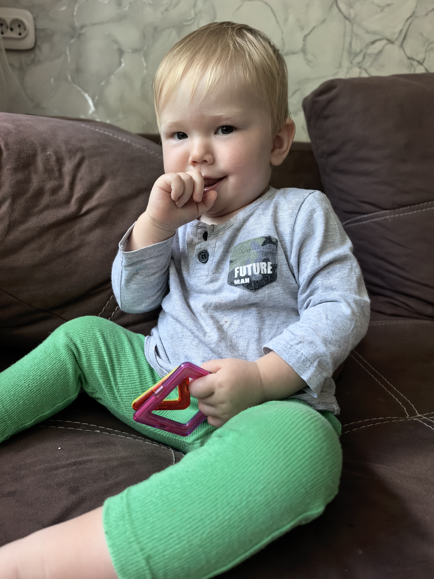 A young, fair-haired child with blue eyes is sitting on a brown couch, placing their thumb in their mouth. The child is wearing a light gray long-sleeved shirt with a patch that says "FUTURE MAN" in black lettering against a camouflage background, and bright green corduroy pants. In their right hand, the child holds a colorful magnetic building toy, a square shape made of pink and yellow translucent plastic. The background consists of a textured wallpaper and a portion of the couch. The image is taken from a medium shot, with the child as the central subject. The lighting is natural, suggesting an indoor environment, possibly a living room. The photograph is taken at an eye-level perspective. There is no information available that indicates the location of this image within the city of Sterlitamak, Russia.