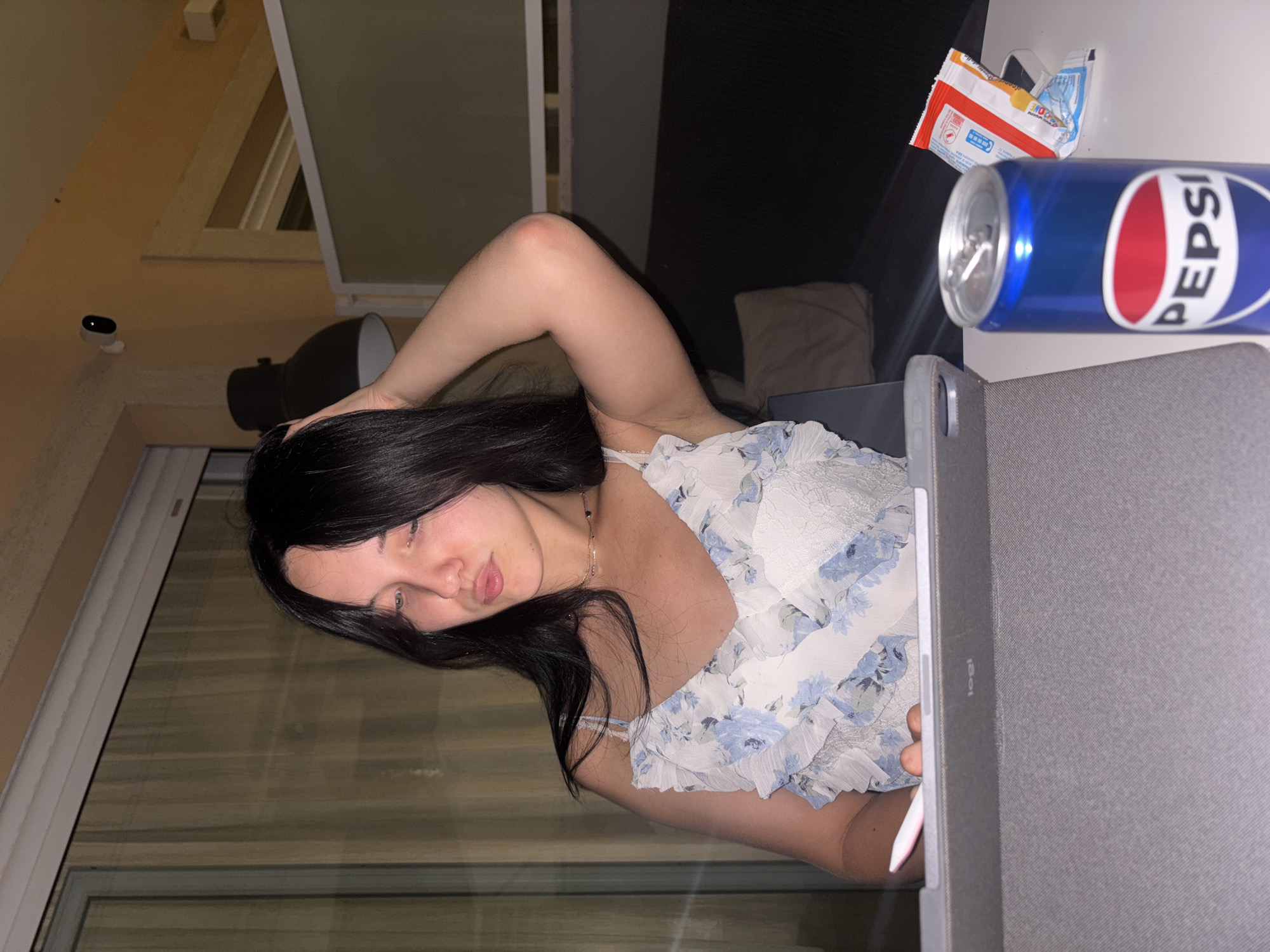 In Cupra Marittima, Italy, a young woman with long, dark hair is seated at a table, her hand in her hair as she looks down. She is wearing a white and blue floral dress. In front of her is a tablet device with a stylus resting on it. To her right, a can of Pepsi and a bag of snacks are placed on the table. The background suggests an indoor setting with a window covered by blinds, and a sofa is visible in the distance. A security camera is mounted on the wall above.