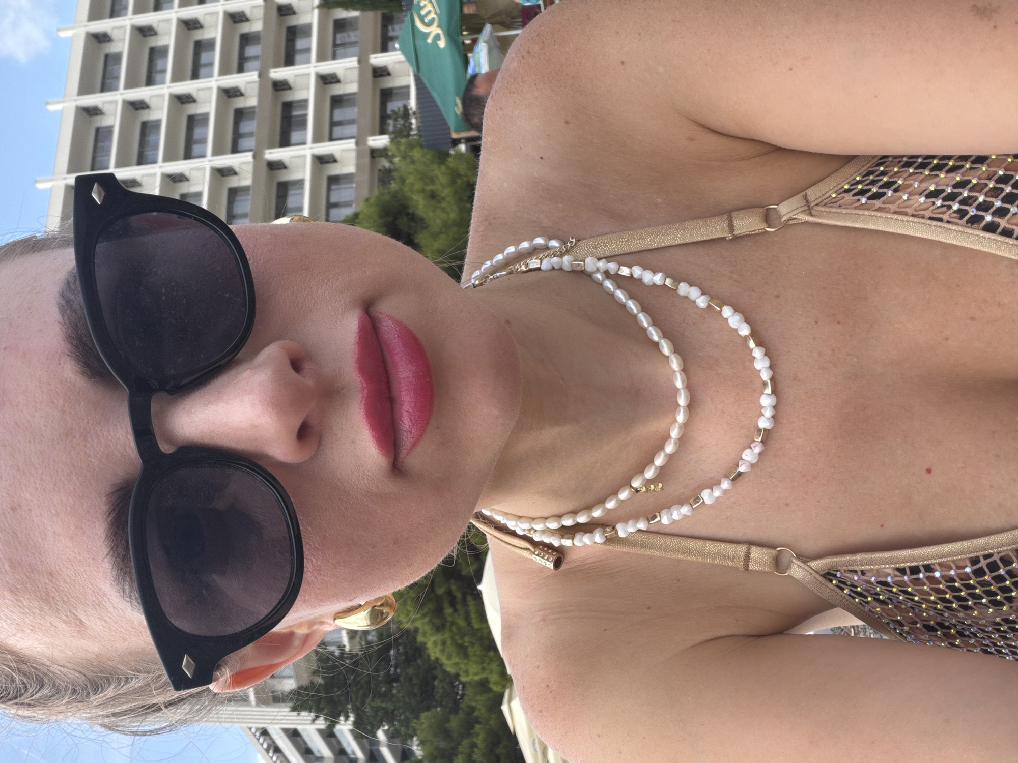The image shows a close-up of a person wearing a gold bikini and sunglasses, with a string of pearls around their neck. The person's face is partially visible, with bright pink lipstick on their lips. In the background, a large white building with many windows is visible, along with some green trees and a blue sky. The image appears to be taken during the day, likely on a sunny day. The setting suggests a vacation or beach location. The overall mood is relaxed and summery. There is no visible text in the image.